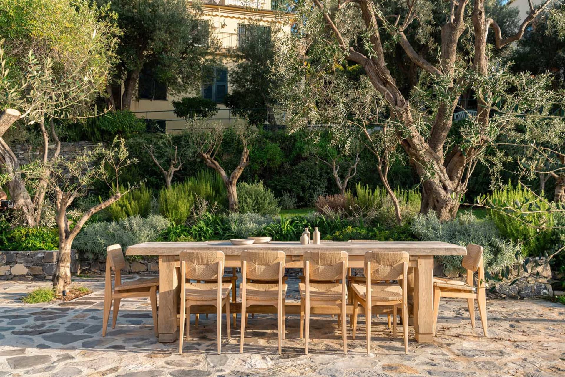 A wooden dining table and chairs set up on a patio surrounded by potted plants, ready for a meal, with a warm and inviting atmosphere