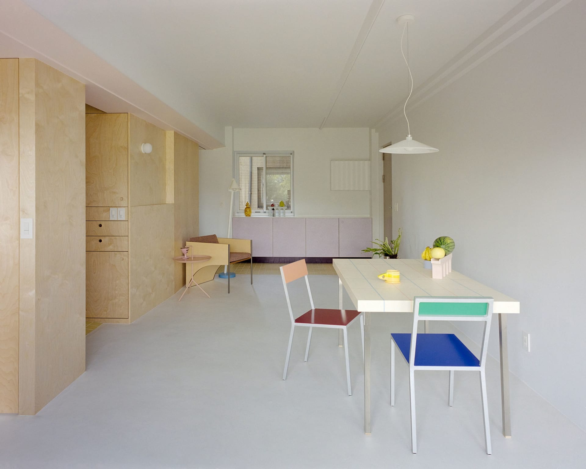 A modern kitchen and dining area with white surfaces, a sink, refrigerator, dining table, chairs, and potted plant, lit by natural sunlight