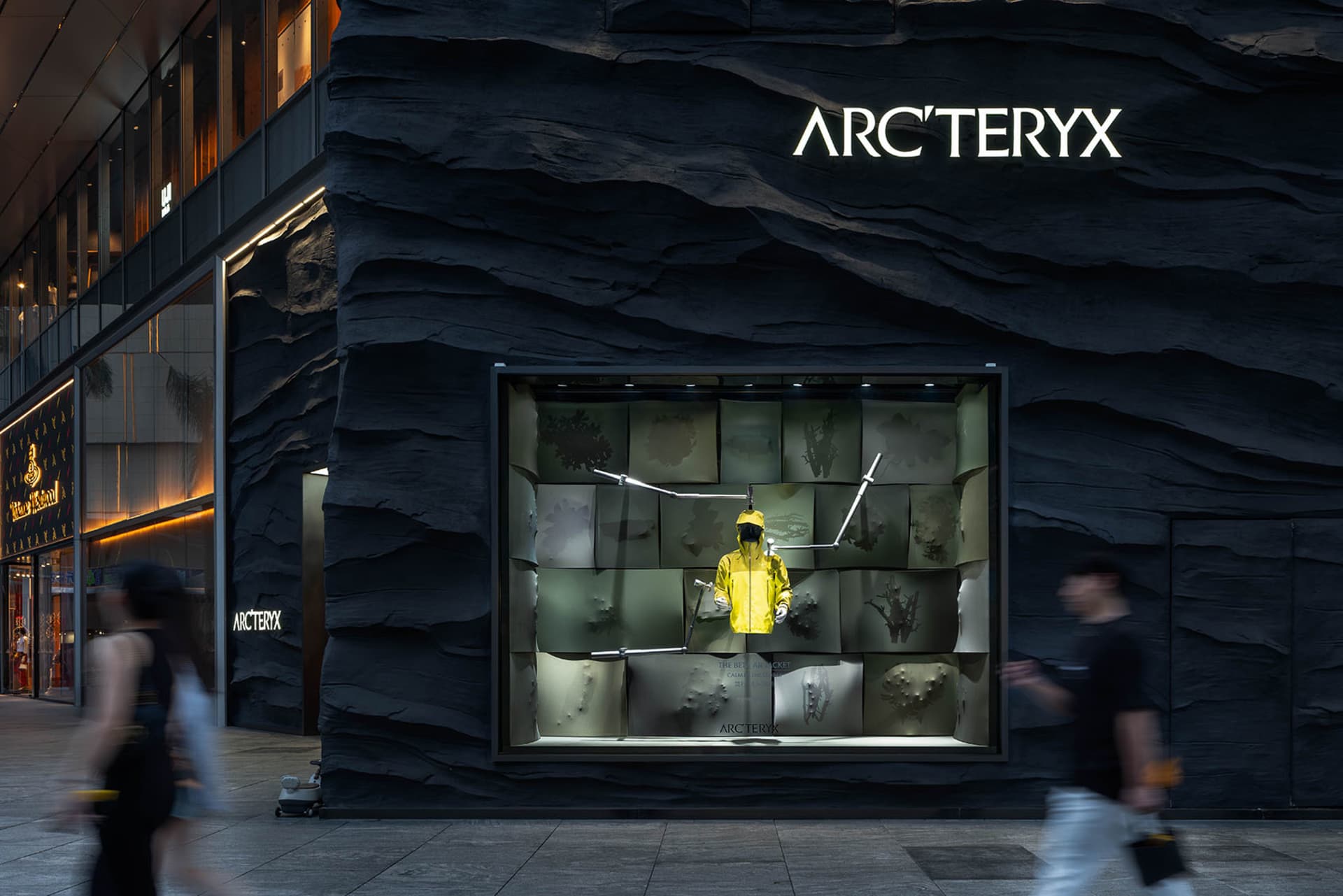 A dimly lit storefront with a stone exterior, featuring a display window with a mannequin wearing a yellow jacket and showcasing various items