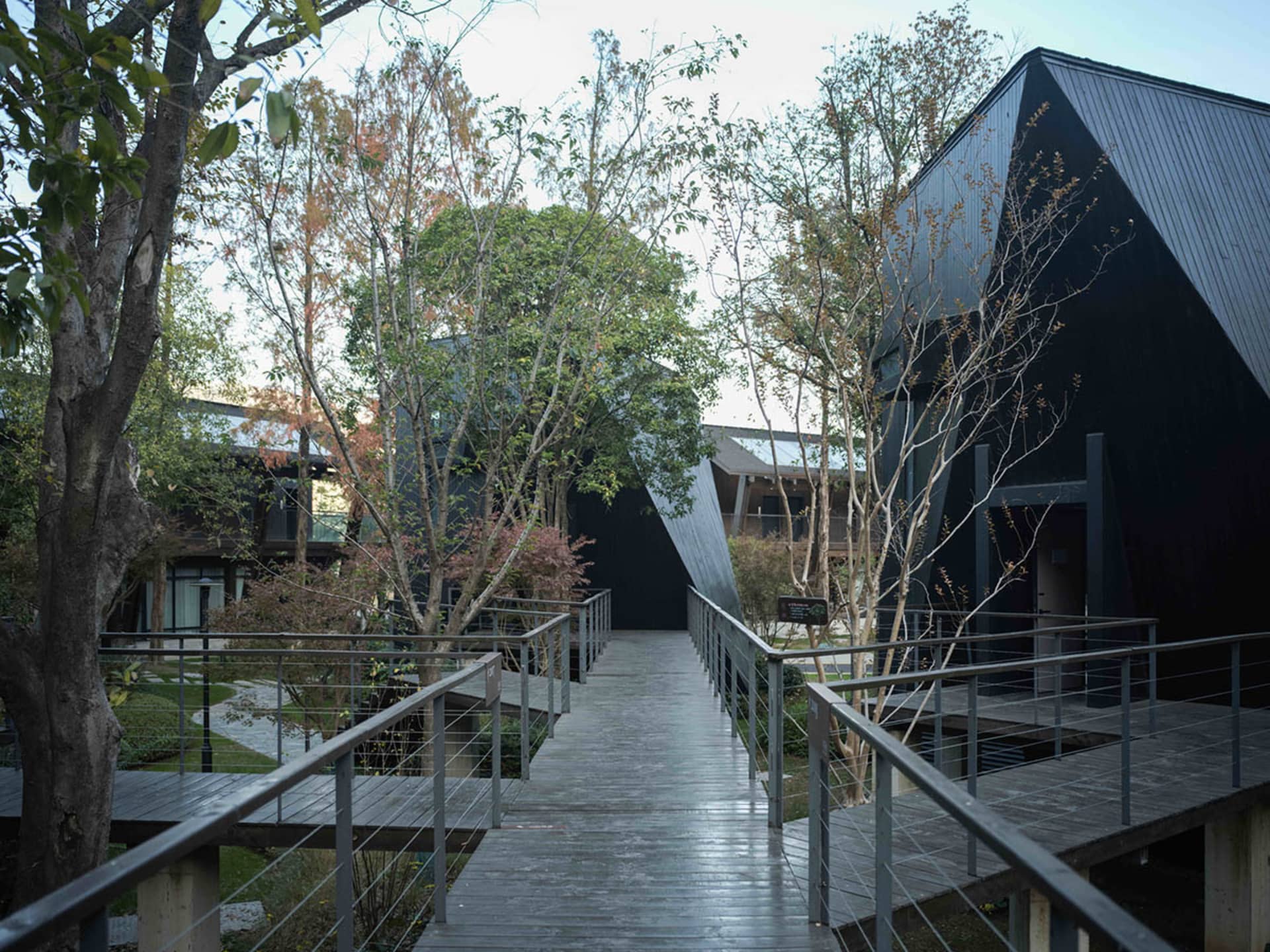 A modern black house with a wooden walkway and metal railing, surrounded by trees, with natural sunlight and a bright atmosphere