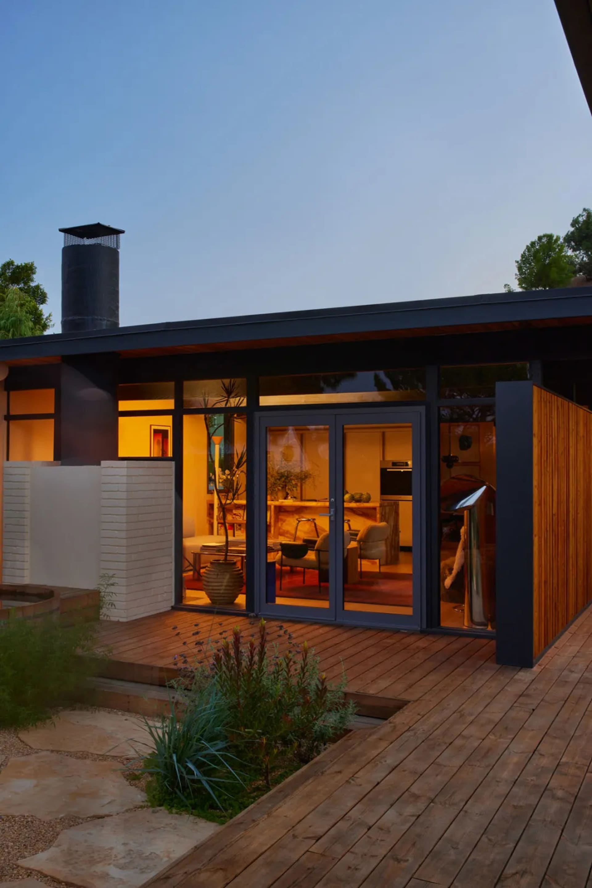 A modern house with a large open living room and kitchen, featuring a fireplace, couch, dining table, and refrigerator, with a black exterior and wooden deck, well-lit for a warm atmosphere