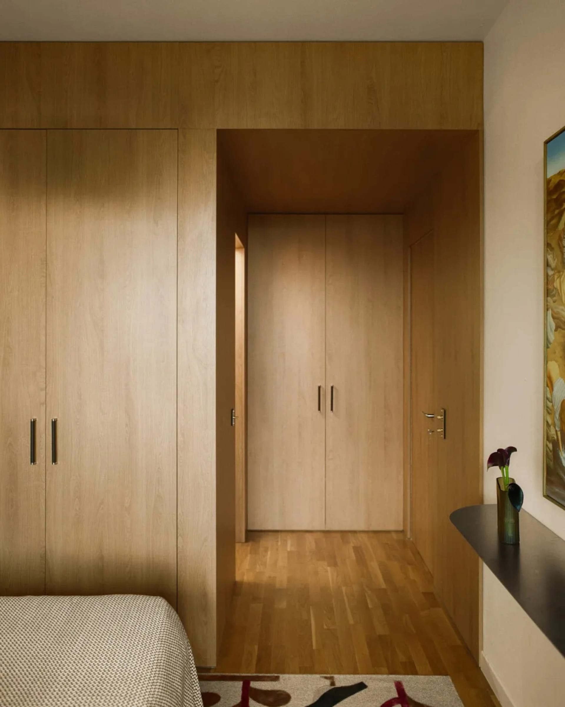 A cozy bedroom with wooden floors and door, featuring a bed, dresser, and painting on the wall, with a warm and natural ambiance
