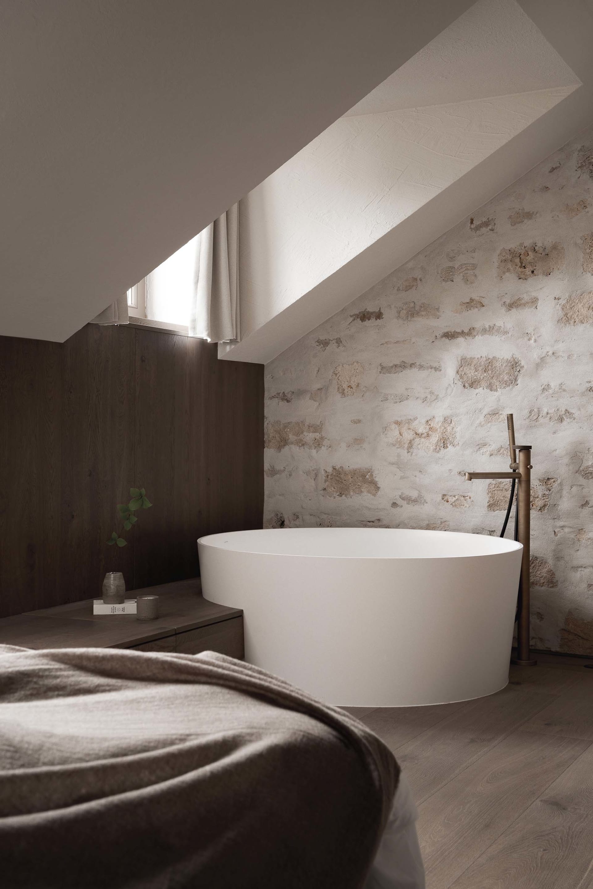 Small bathroom with a white bathtub, sink, and window, set against a stone wall and wooden floor in a dimly lit space