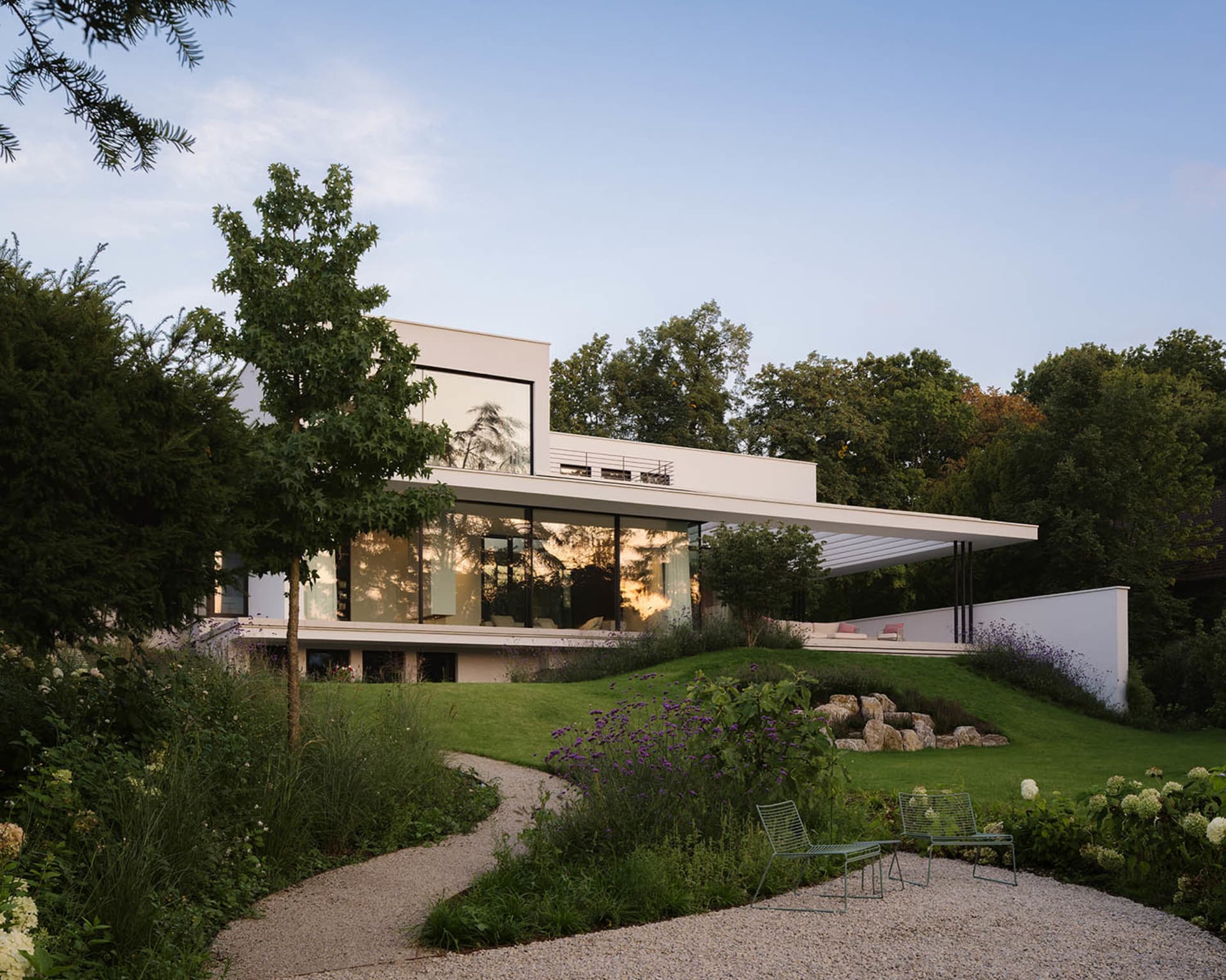 A serene residential house with a white exterior, large window, and patio area, surrounded by a garden with a tree and bushes
