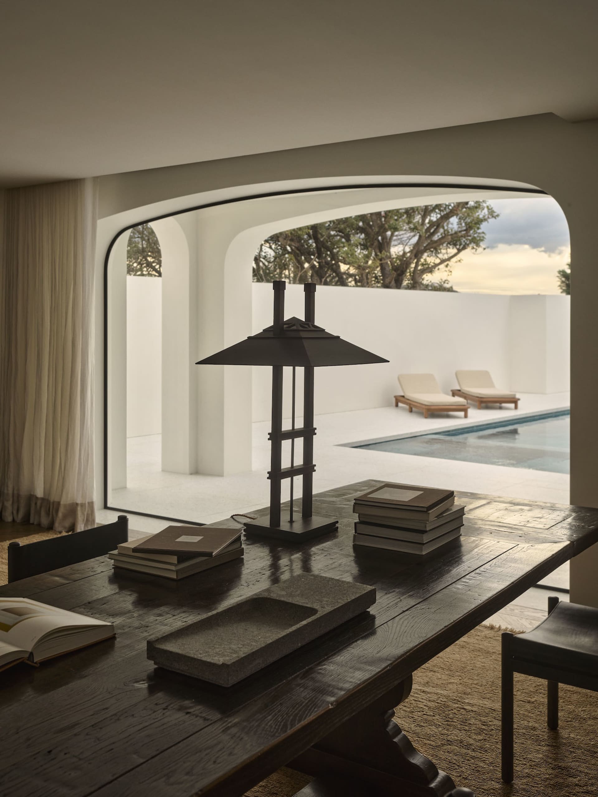 A modern living room with a pool in the background, featuring a dining table, two couches, and various books, creating a cozy and inviting atmosphere