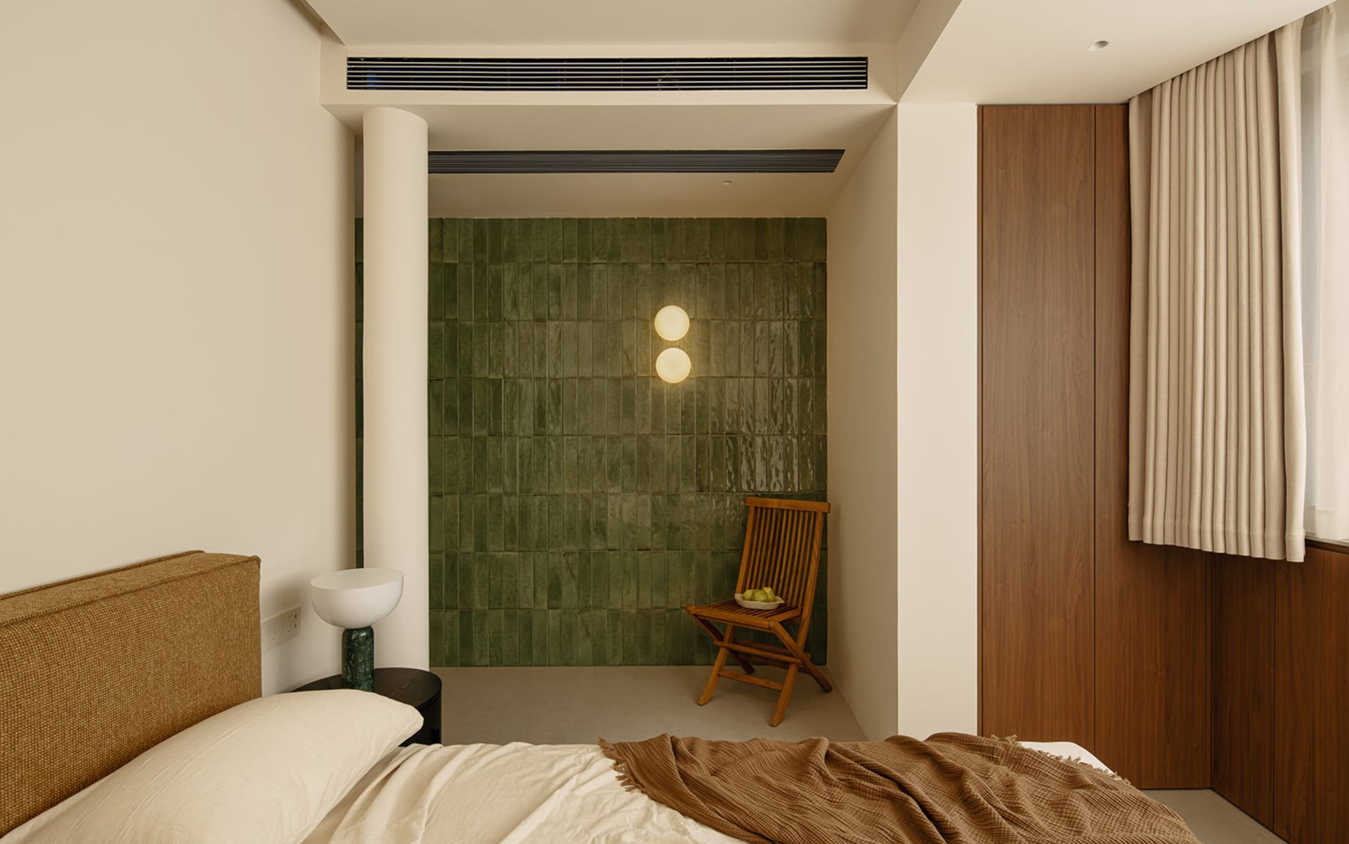 A modern bedroom with a green tiled wall, white floor, and a bed, chair, and light, illuminated by natural sunlight through a window