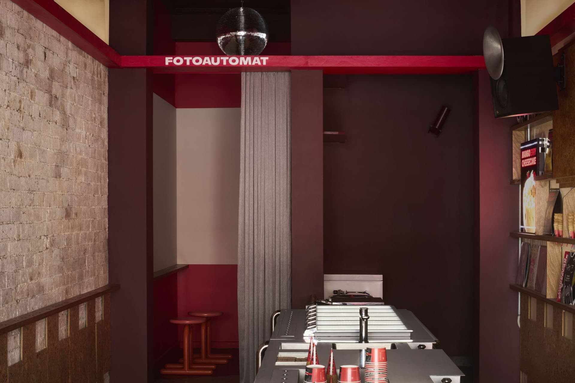 A small, dark room with a red wall, a black counter, and a few chairs, featuring cups and bottles on the counter