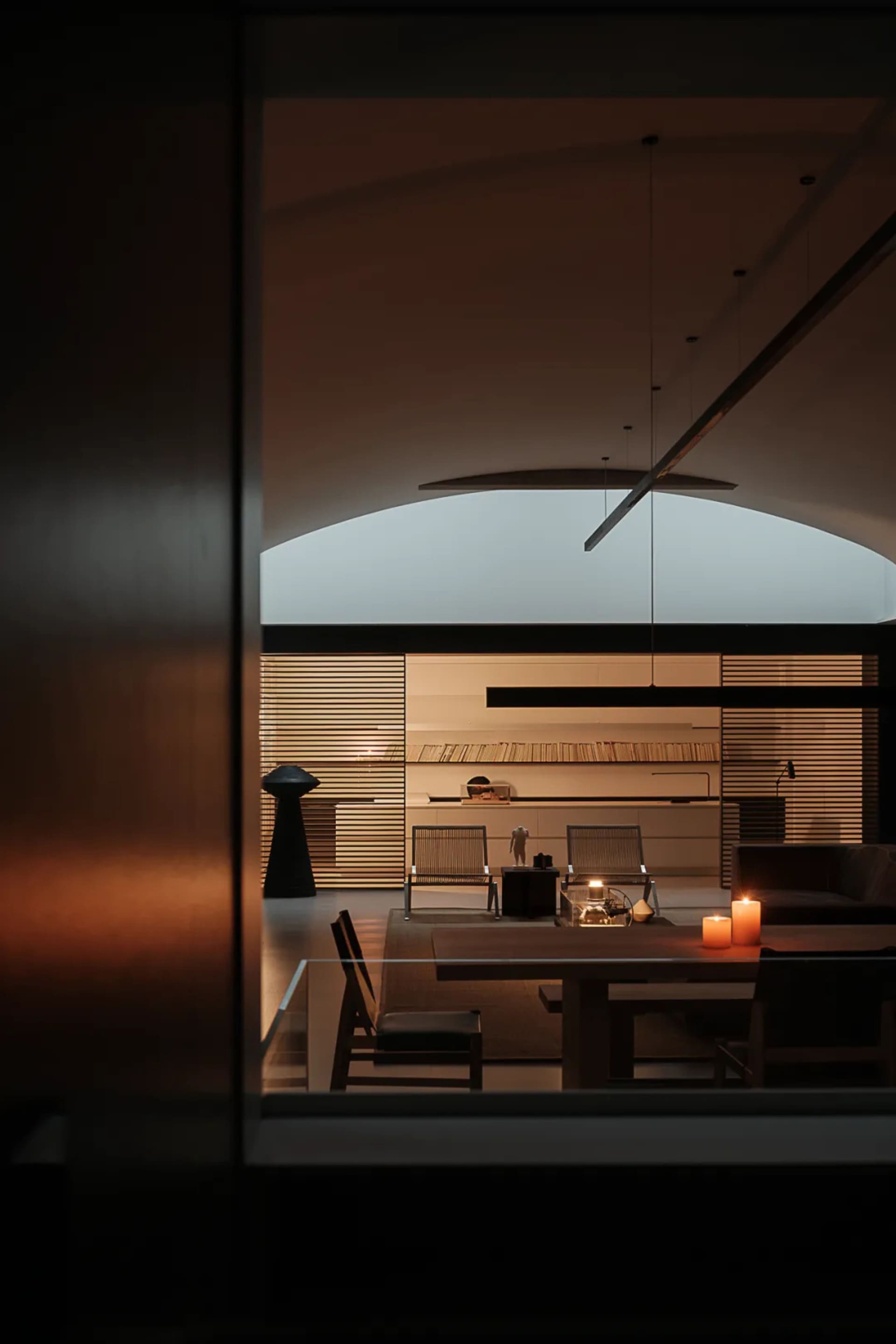 A modern dining room with a large window, a dining table, chairs, a couch, and a bookshelf, lit by natural light and a candle, creating a warm and inviting atmosphere