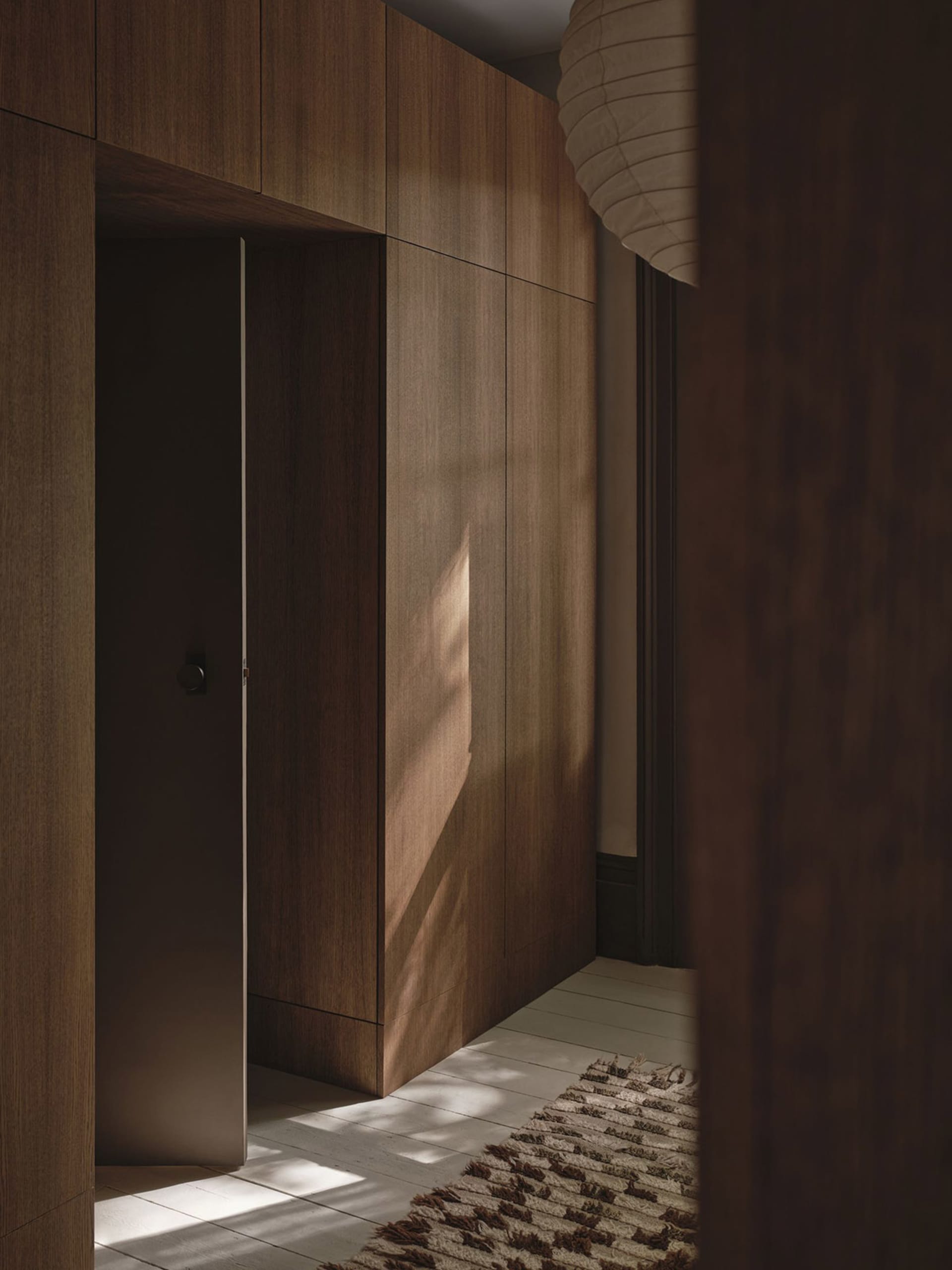 A dimly lit hallway with a wooden door, wooden cabinet, and wooden floor, with a doorway leading to another room