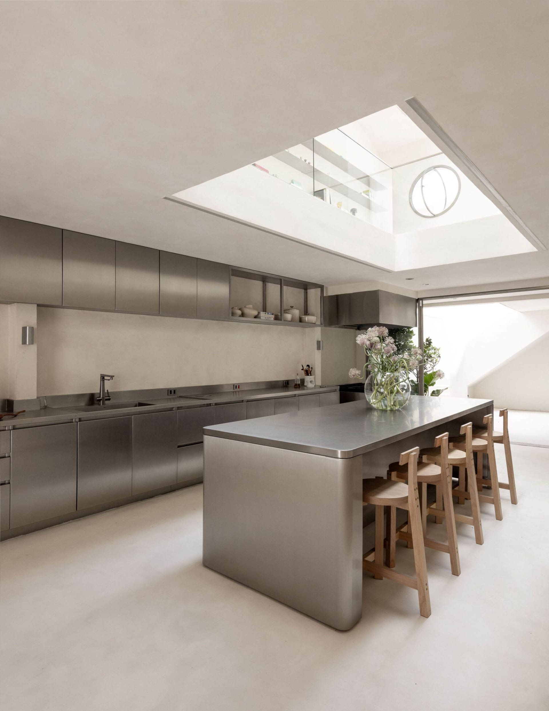 A well-lit kitchen with a large steel island, wooden chairs, and a potted plant, surrounded by white walls and a steel sink