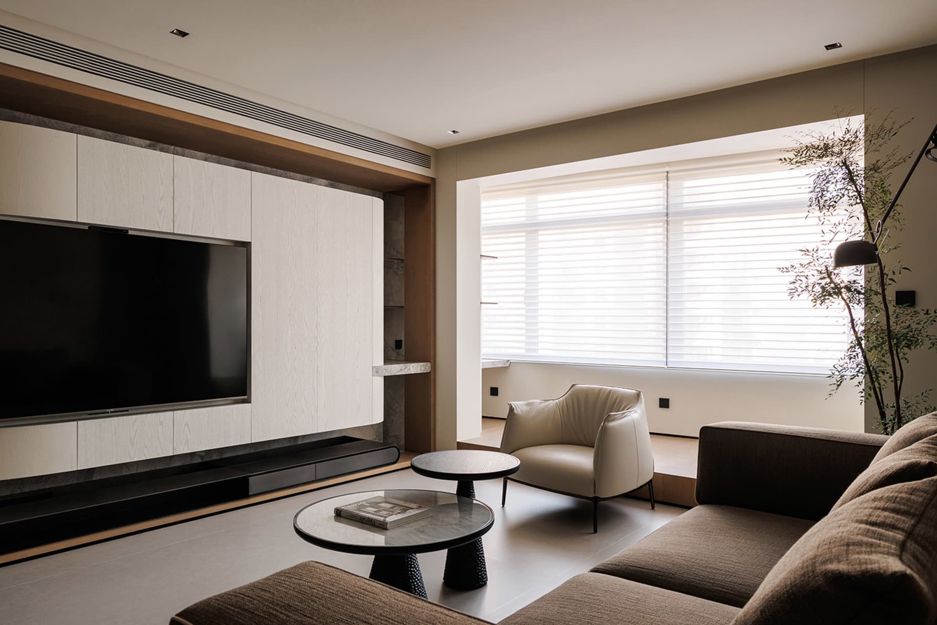 Cozy modern living space with a large flat screen TV, comfortable couch, chair, and dining table, decorated with books, a vase, and a remote control, with natural light from the windows