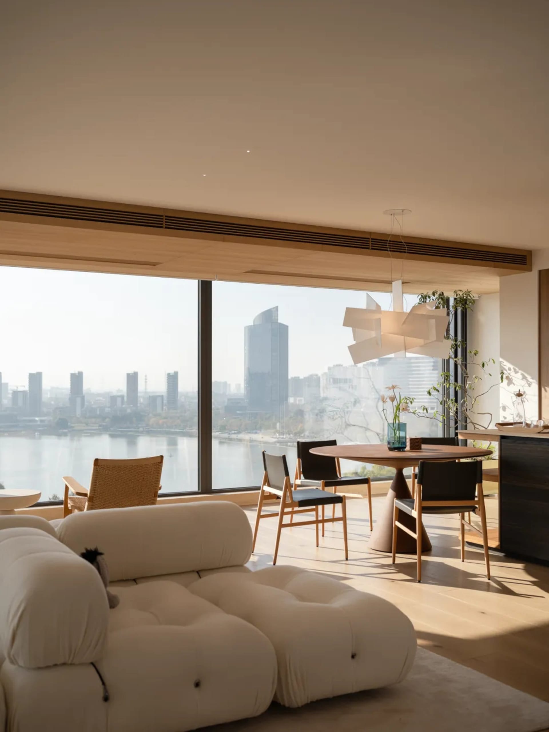 Modern living room with a large window, couch, dining table, chairs, potted plants, and vases, with natural light and a city view