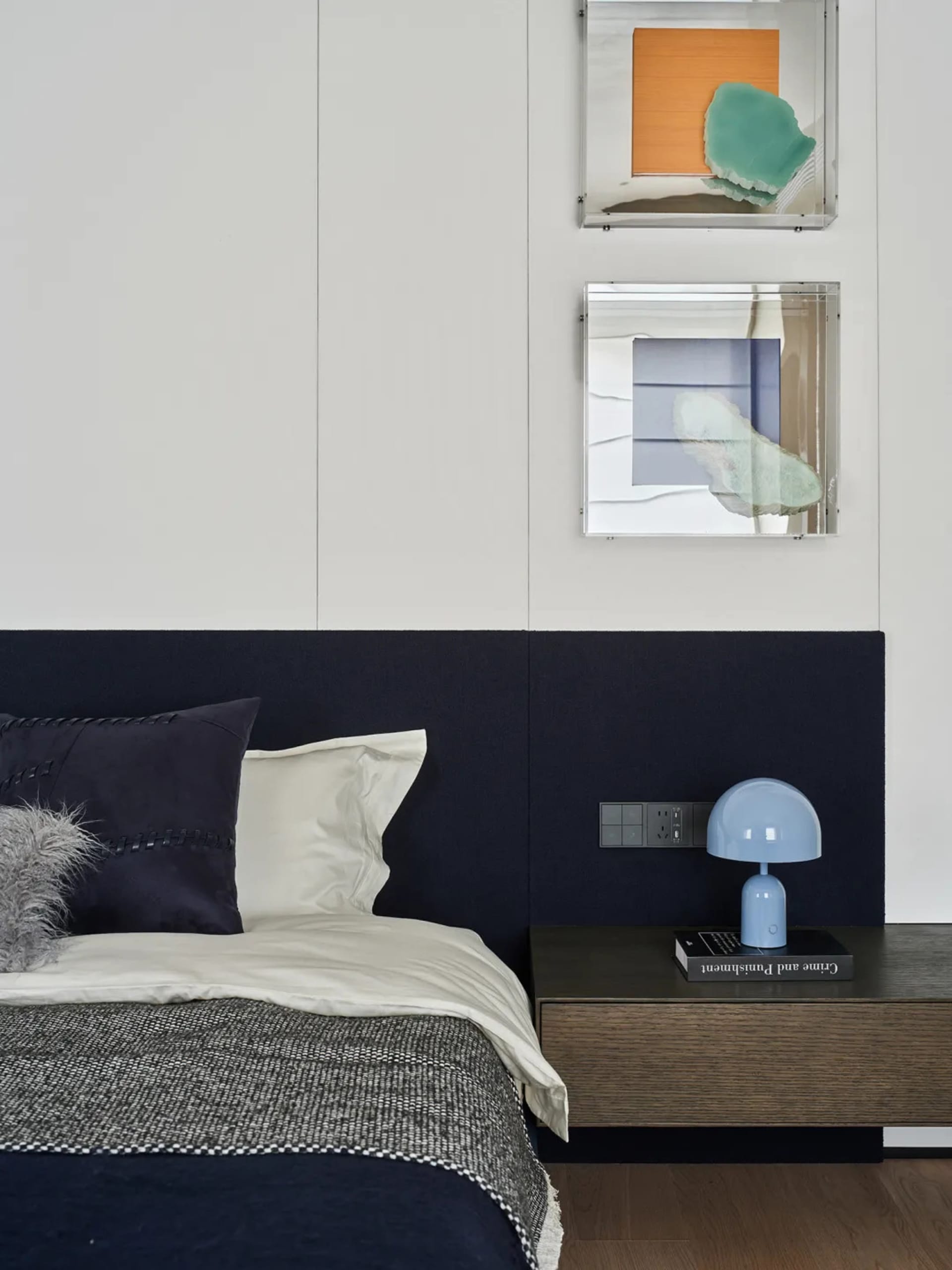 A modern bedroom with a white comforter, a blue lamp, and a row of three pictures on the wall, set against a well-lit background