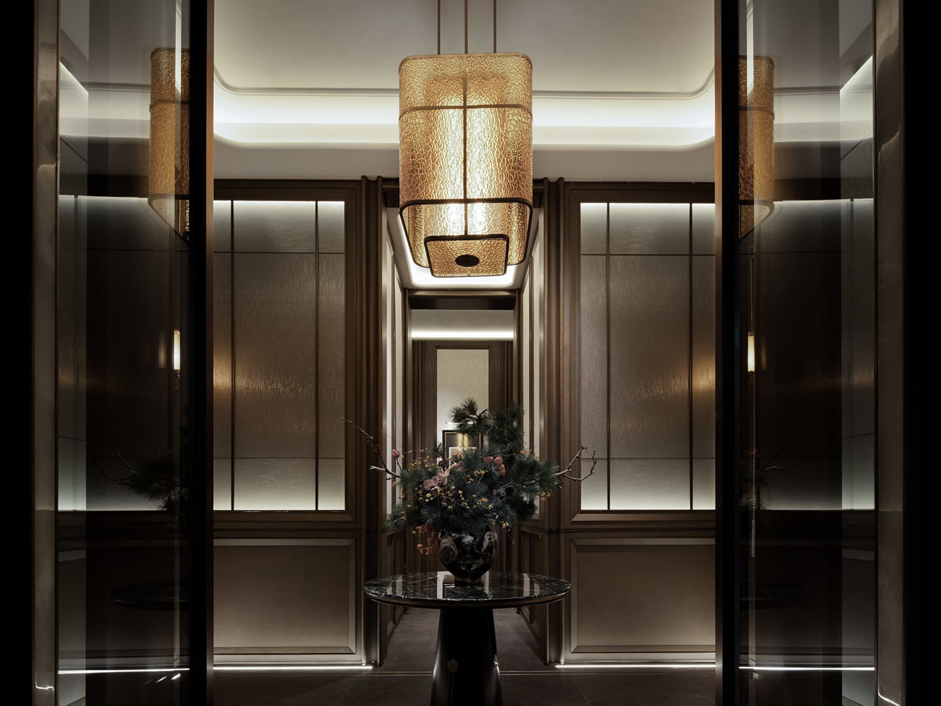 A well-lit hallway with a glass vase on a table and a metal chandelier hanging from the ceiling, surrounded by doors and decorated with flowers