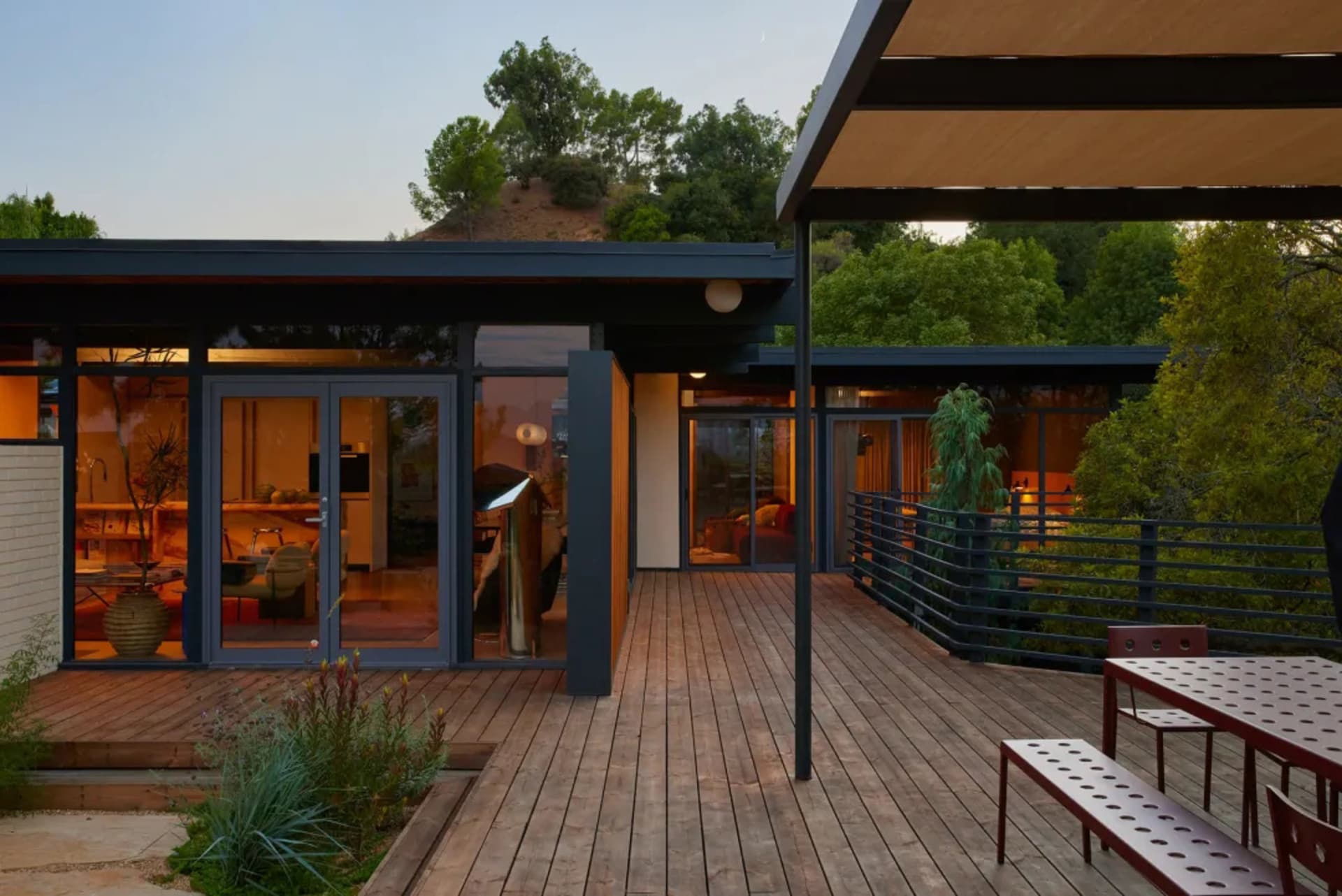 A modern house with a wooden deck and patio surrounded by trees, featuring a large window, TV, and natural sunlight