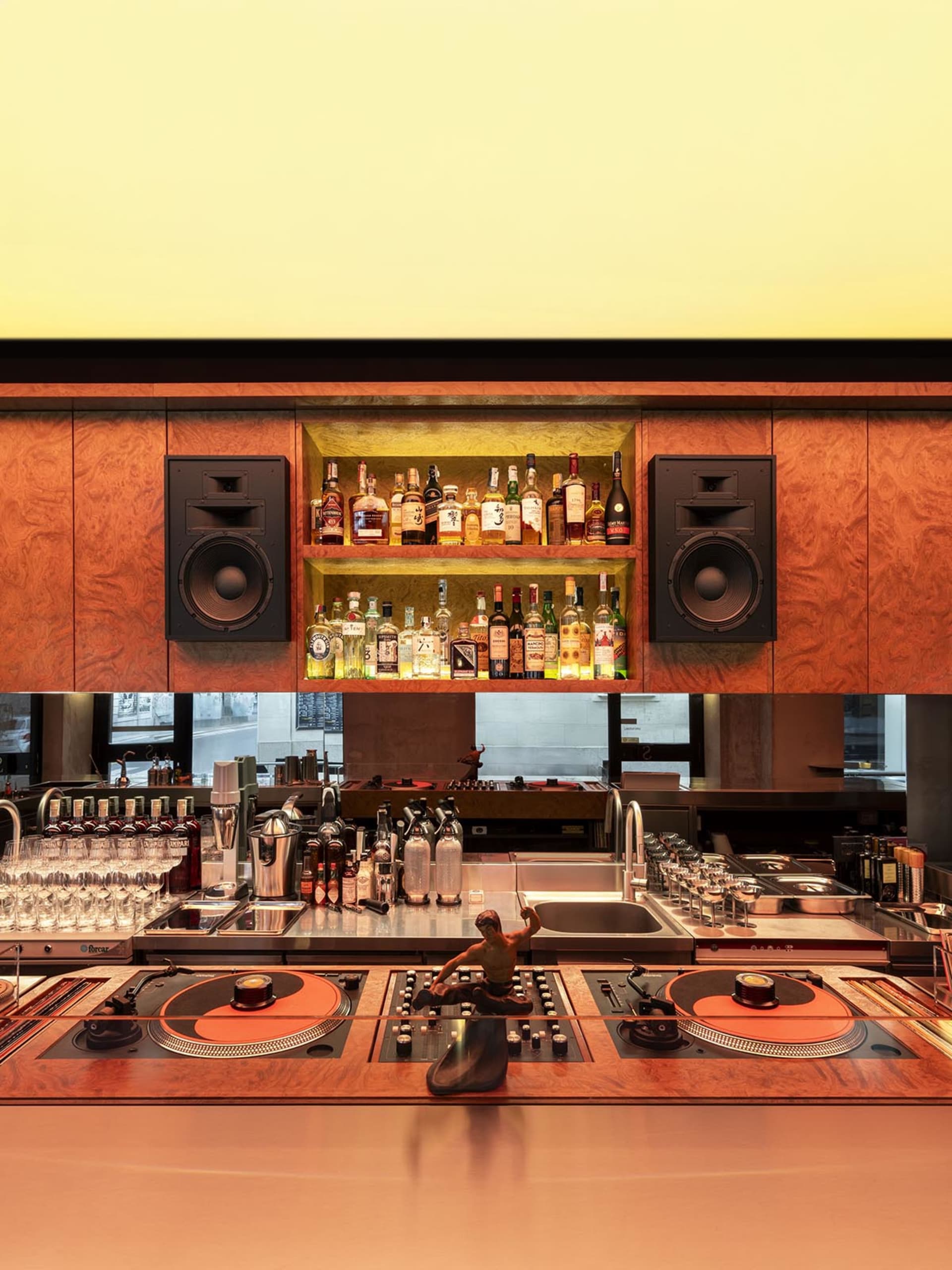 A dimly lit modern bar with a wooden countertop, displaying a variety of liquor bottles and glasses, creating a cozy and inviting atmosphere
