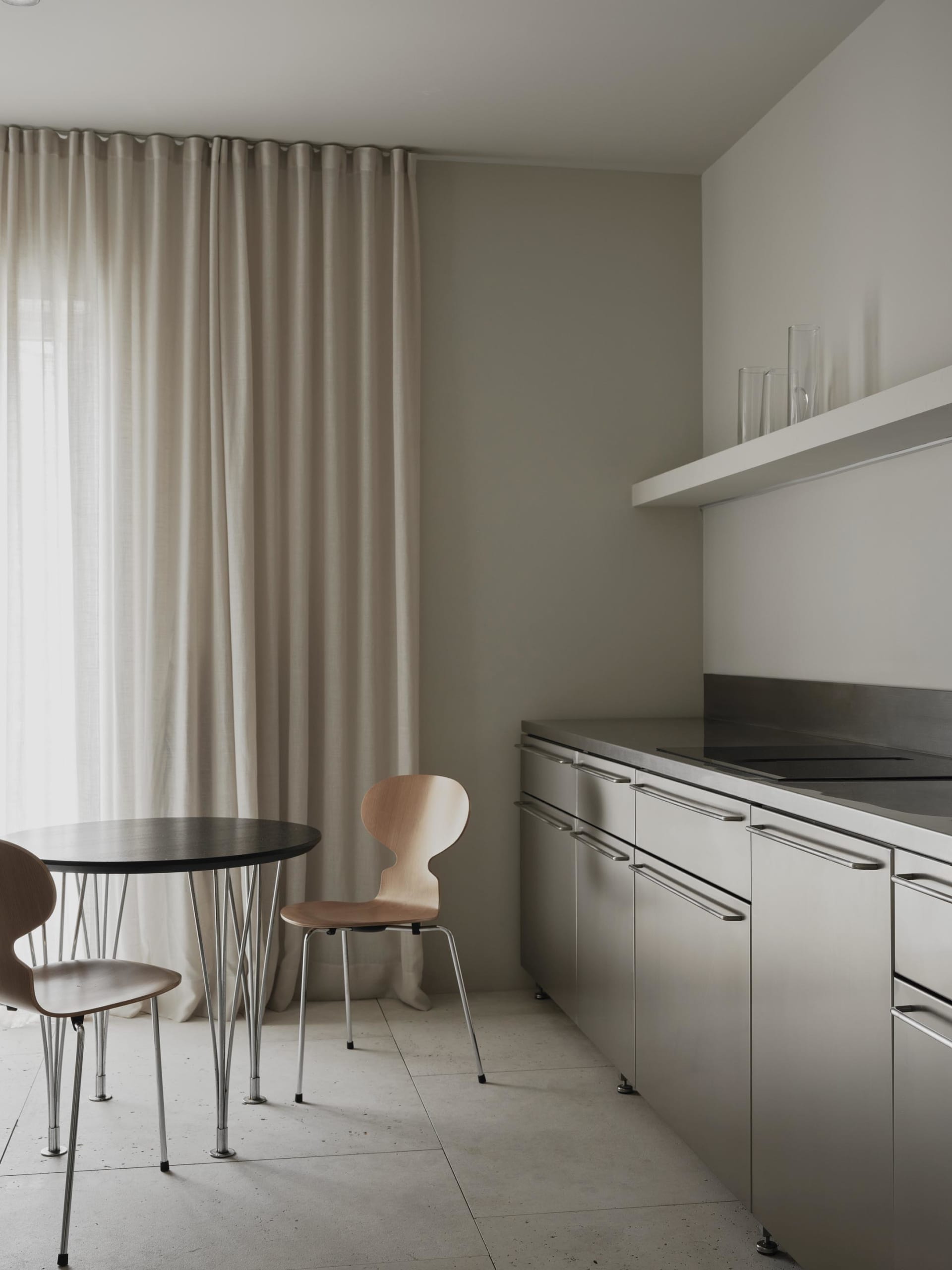 A modern kitchen with a dining table and chairs, surrounded by white surfaces, with natural light and a sink in the background