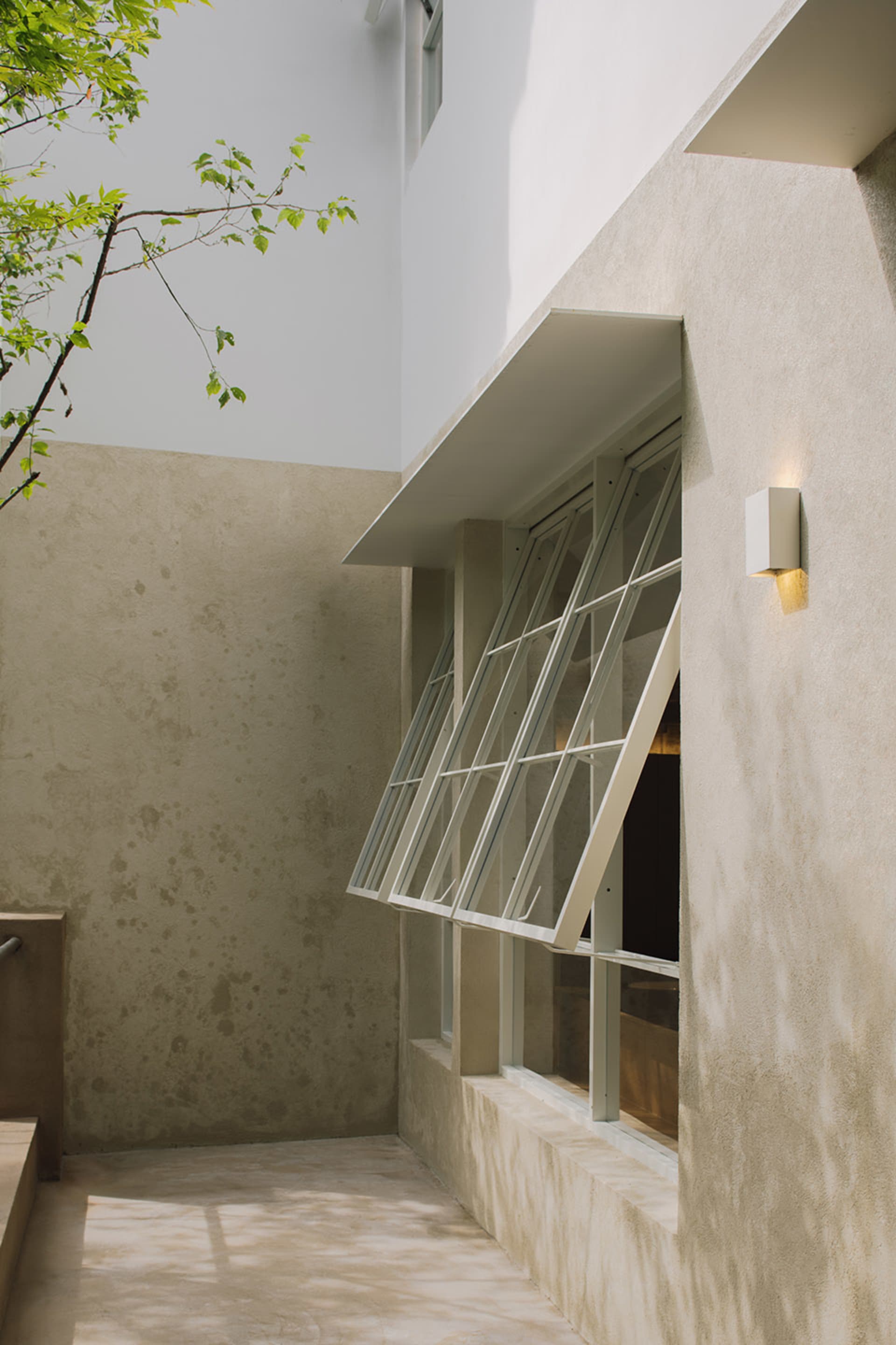A stone building with an open window and closed door, set in a serene courtyard with a tree in the background