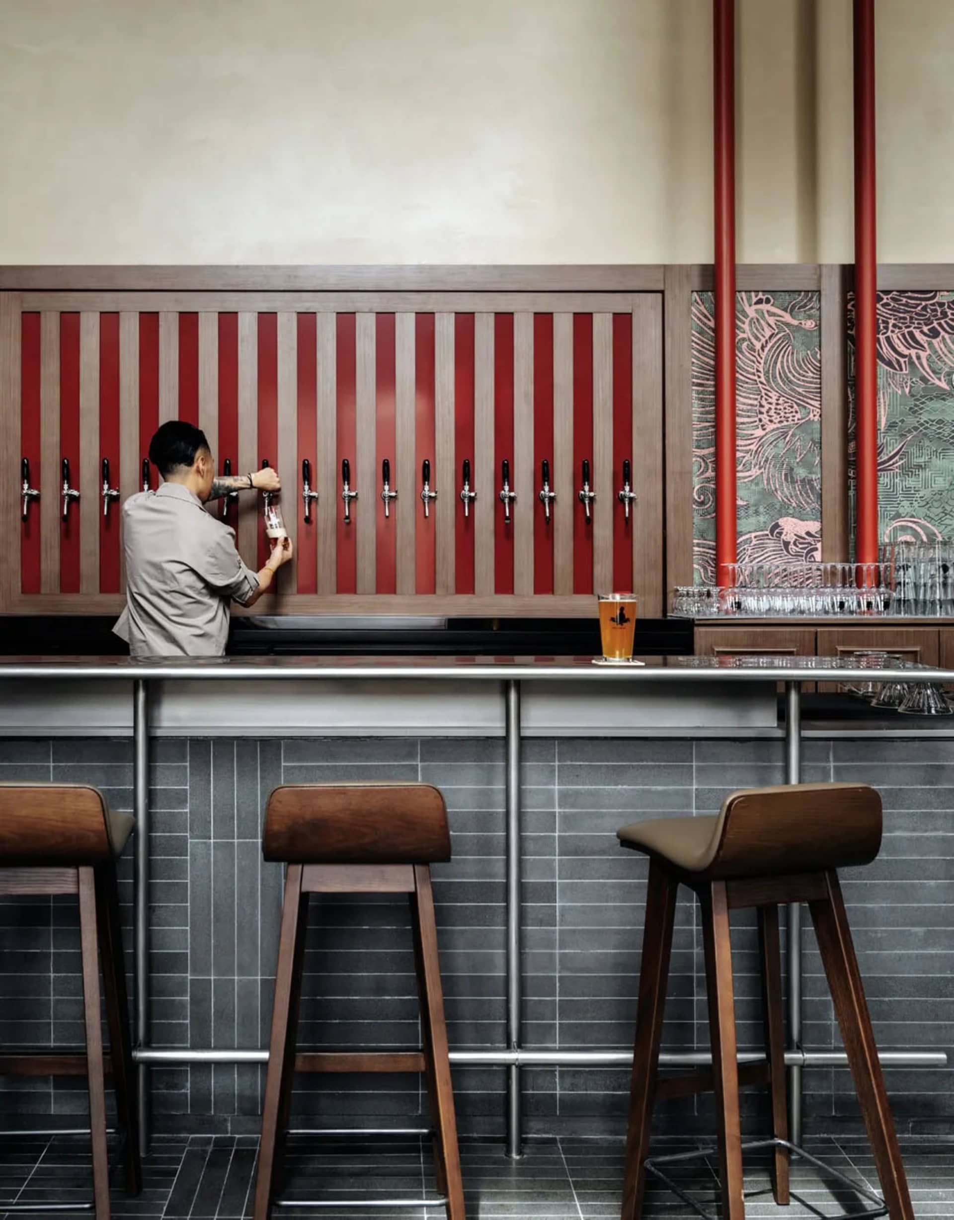 A casual bar scene with a man standing in front of a bar with a red and brown color scheme, beer taps, chairs, and a dining table