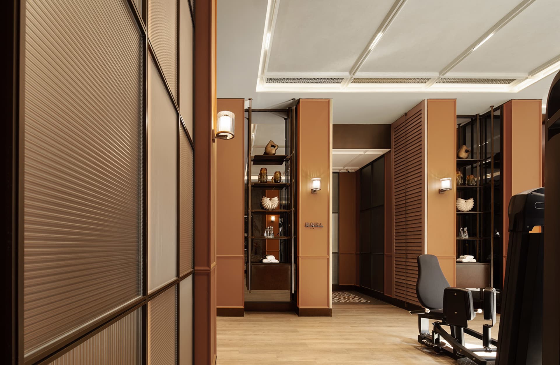 A dimly lit hallway with a wooden floor and brown walls, lined with doors and featuring a chair and bottles on surfaces