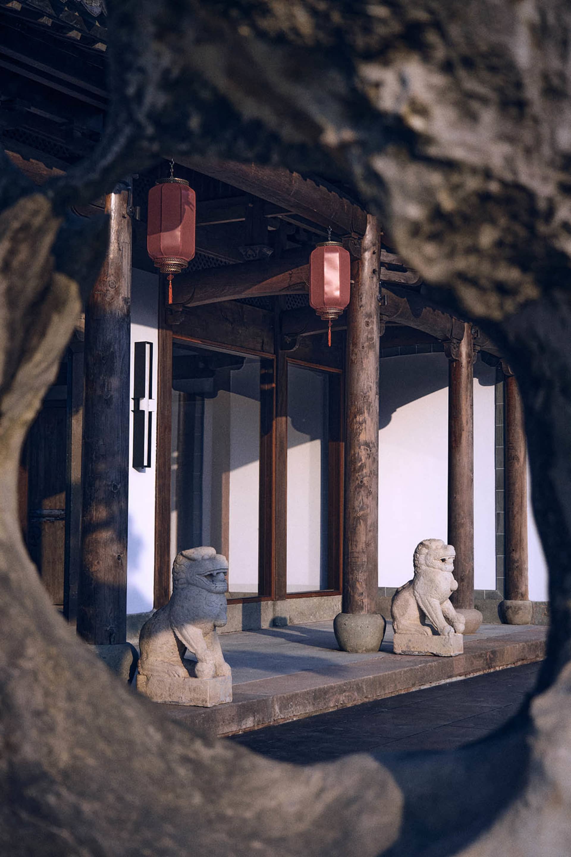 A stone building with a wooden structure, adorned with red lanterns and featuring two lion statues on a stone bench, set against a natural light-filled backdrop