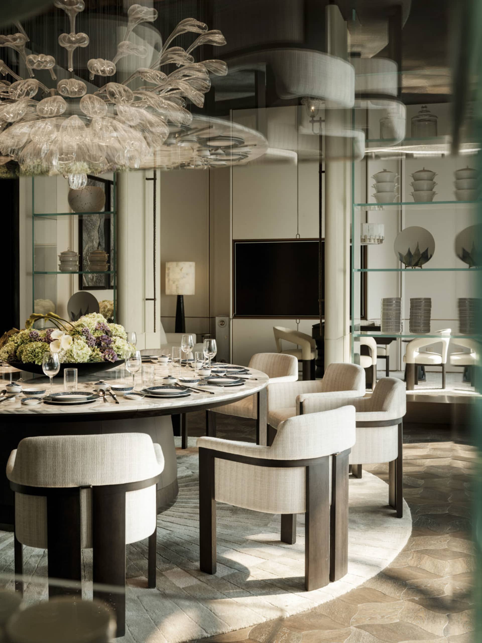 A well-lit dining room with a round table, chairs, vases, and a chandelier, surrounded by wood and glass surfaces