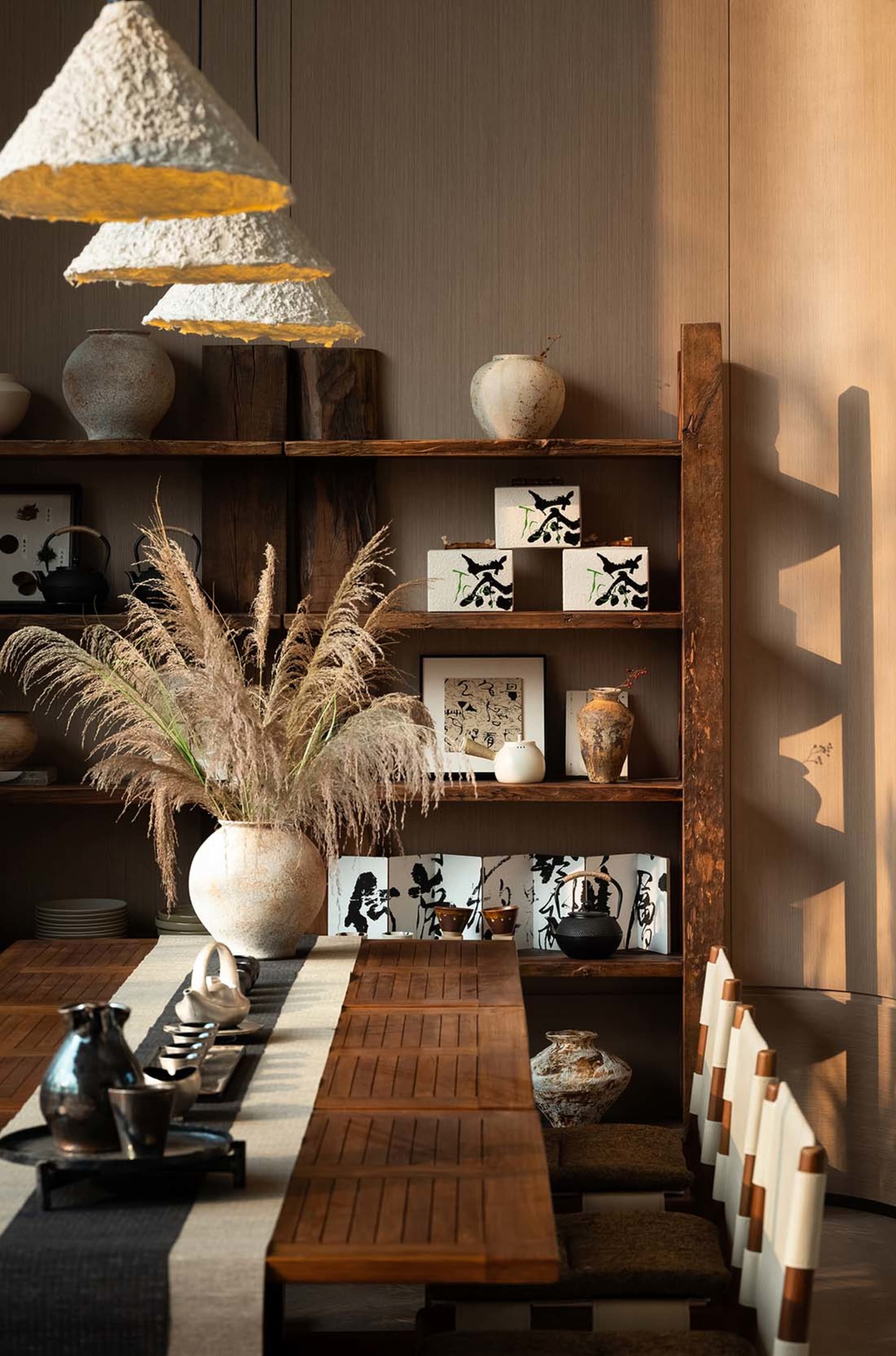 Dining room with a wooden table, chairs, and bookshelf, set with various items and decorated with vases, surrounded by natural light