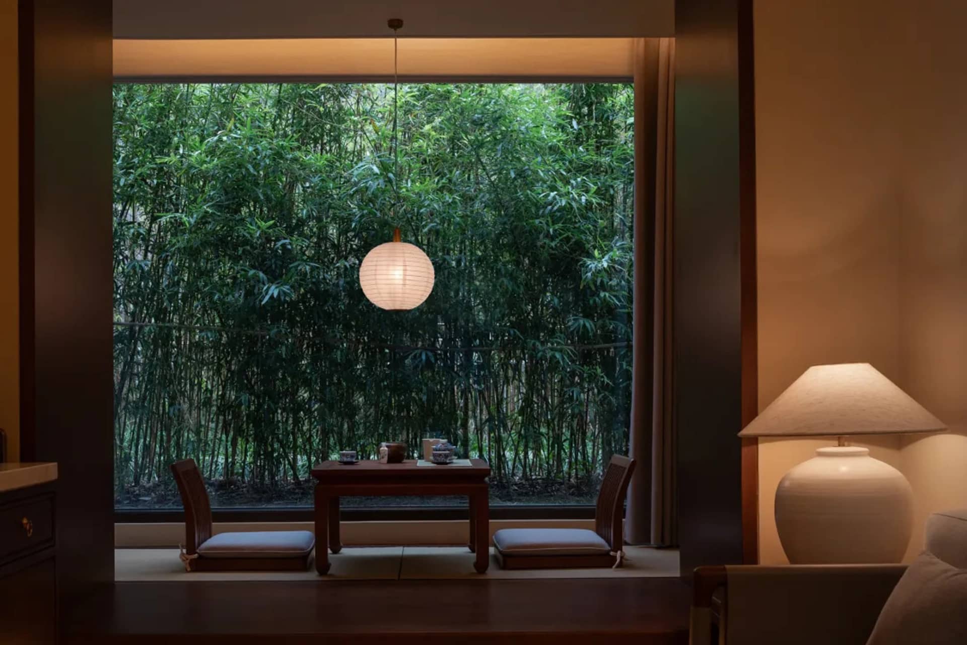 A dining room with a wooden table and chairs, a lamp, and a window with a forest view, creating a warm and inviting atmosphere