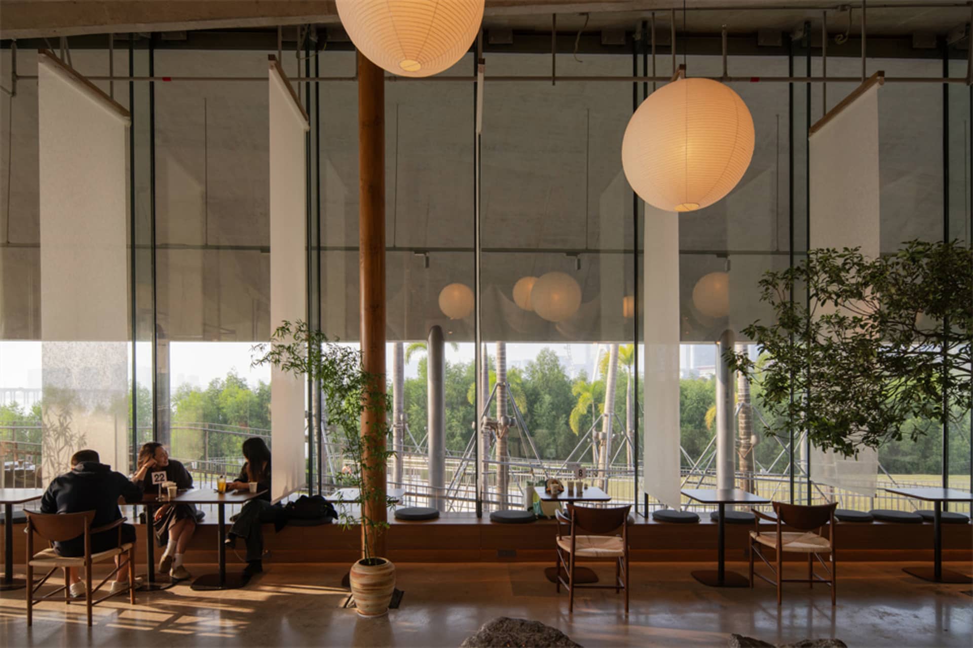 A bright and welcoming restaurant with wooden tables, chairs, and potted plants, flooded with natural light through a large window