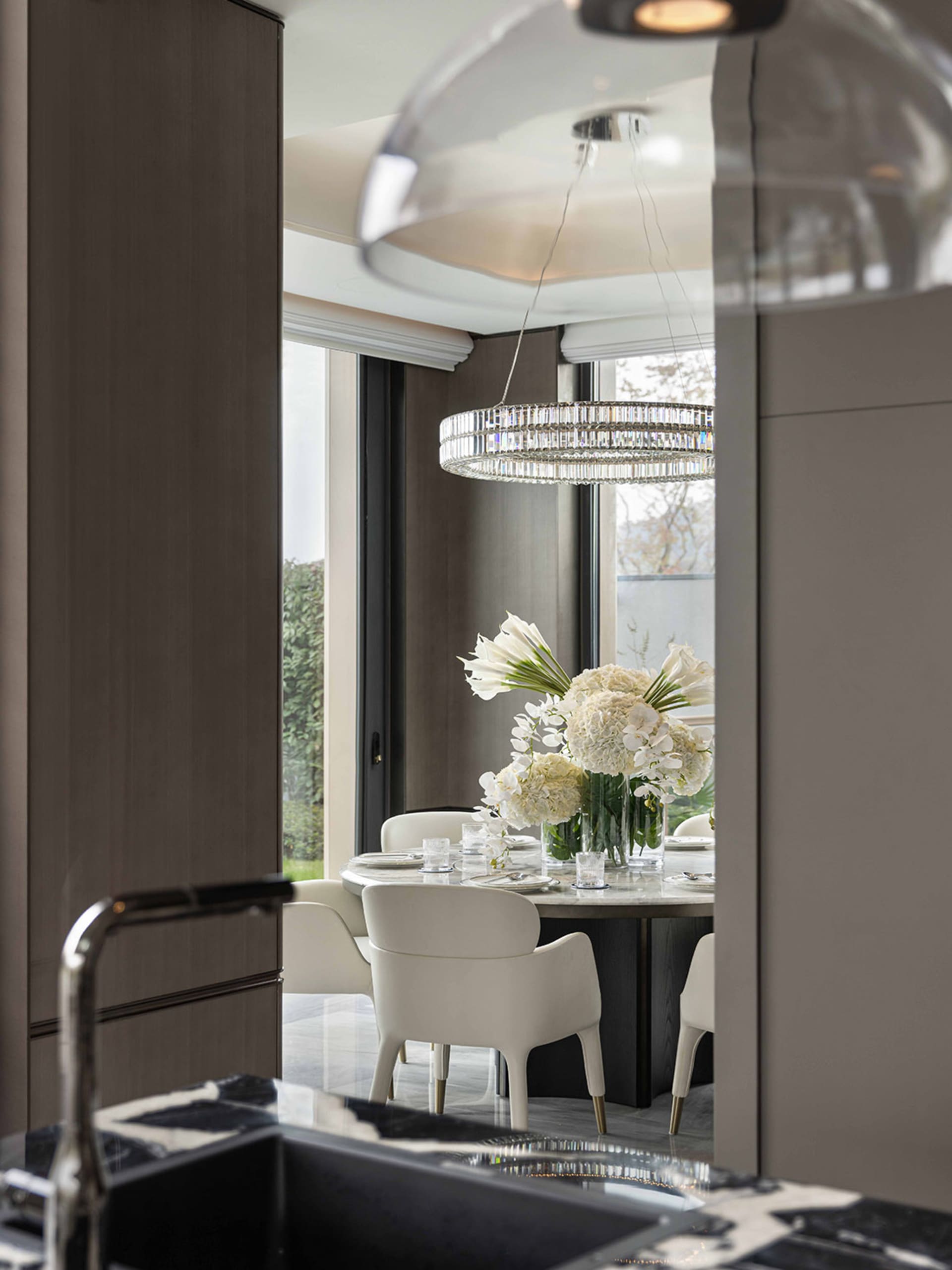 Dining room with a large window, a chandelier, and a table with white chairs, set with a vase of flowers, in a modern space with natural light