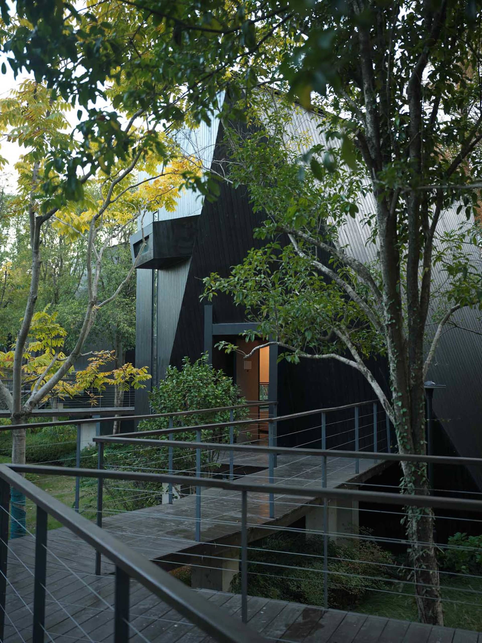 A modern house with a black exterior, a wooden deck, and a metal staircase, surrounded by trees and natural light