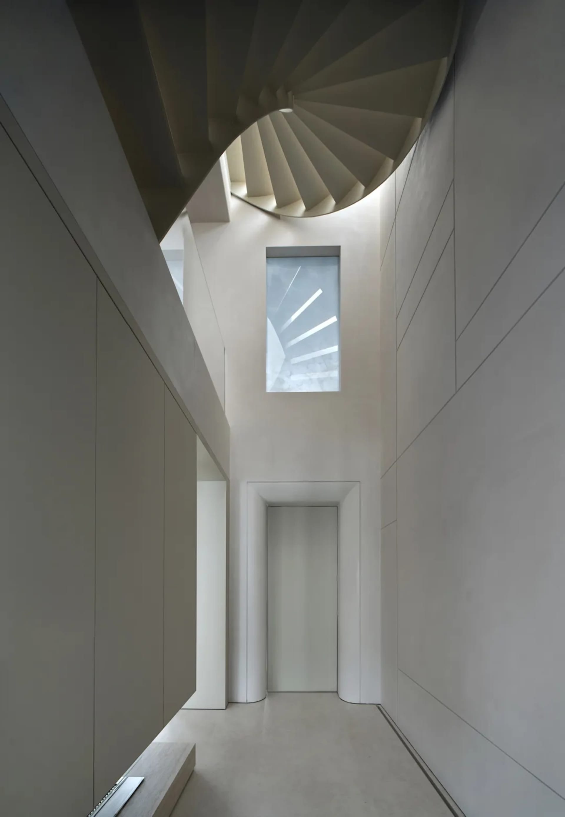 A modern hallway with a concrete spiral staircase, illuminated by natural light from a window, creating a calm and inviting atmosphere