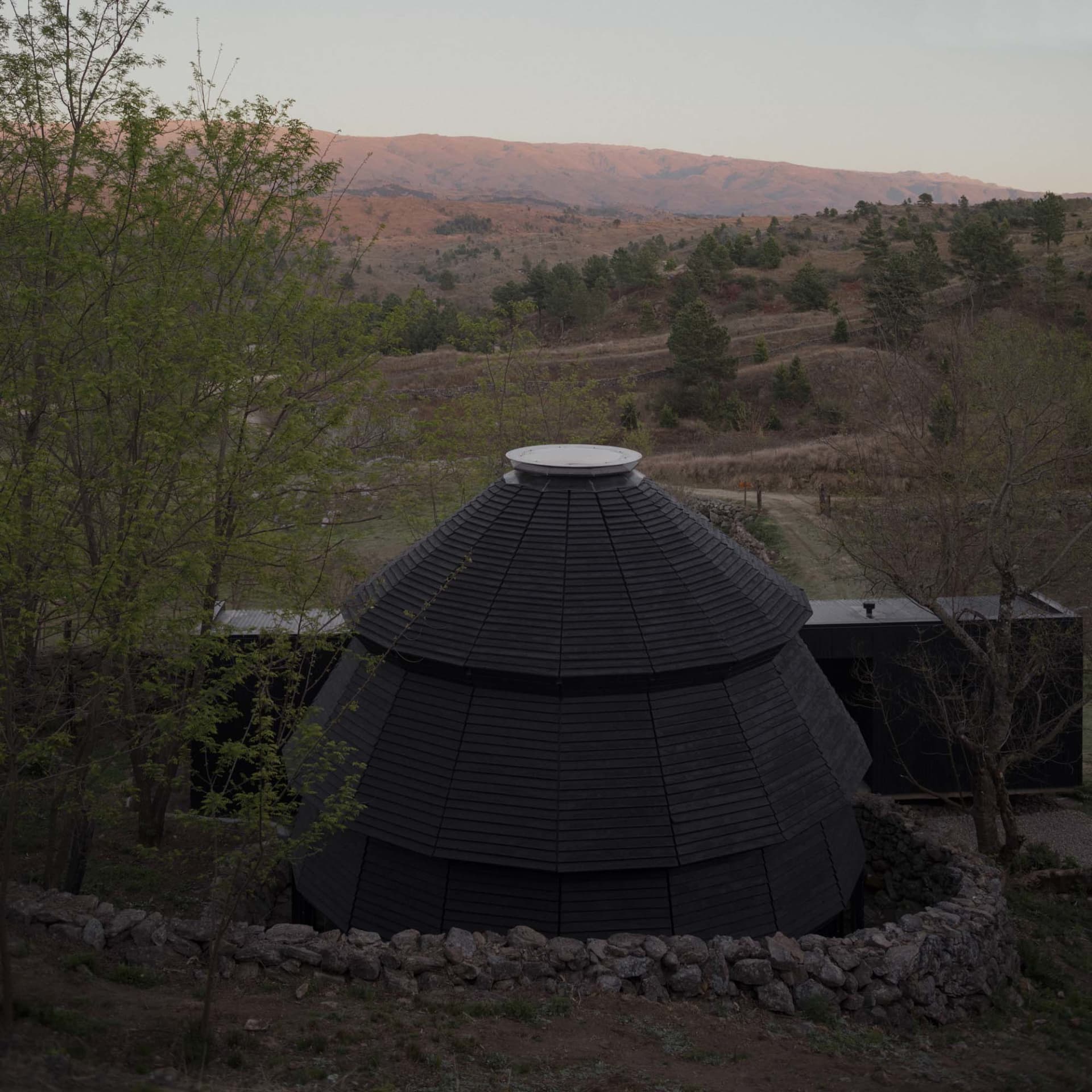 A serene black and white photograph of a unique house with a large stone dome on a hillside, surrounded by trees and mountains