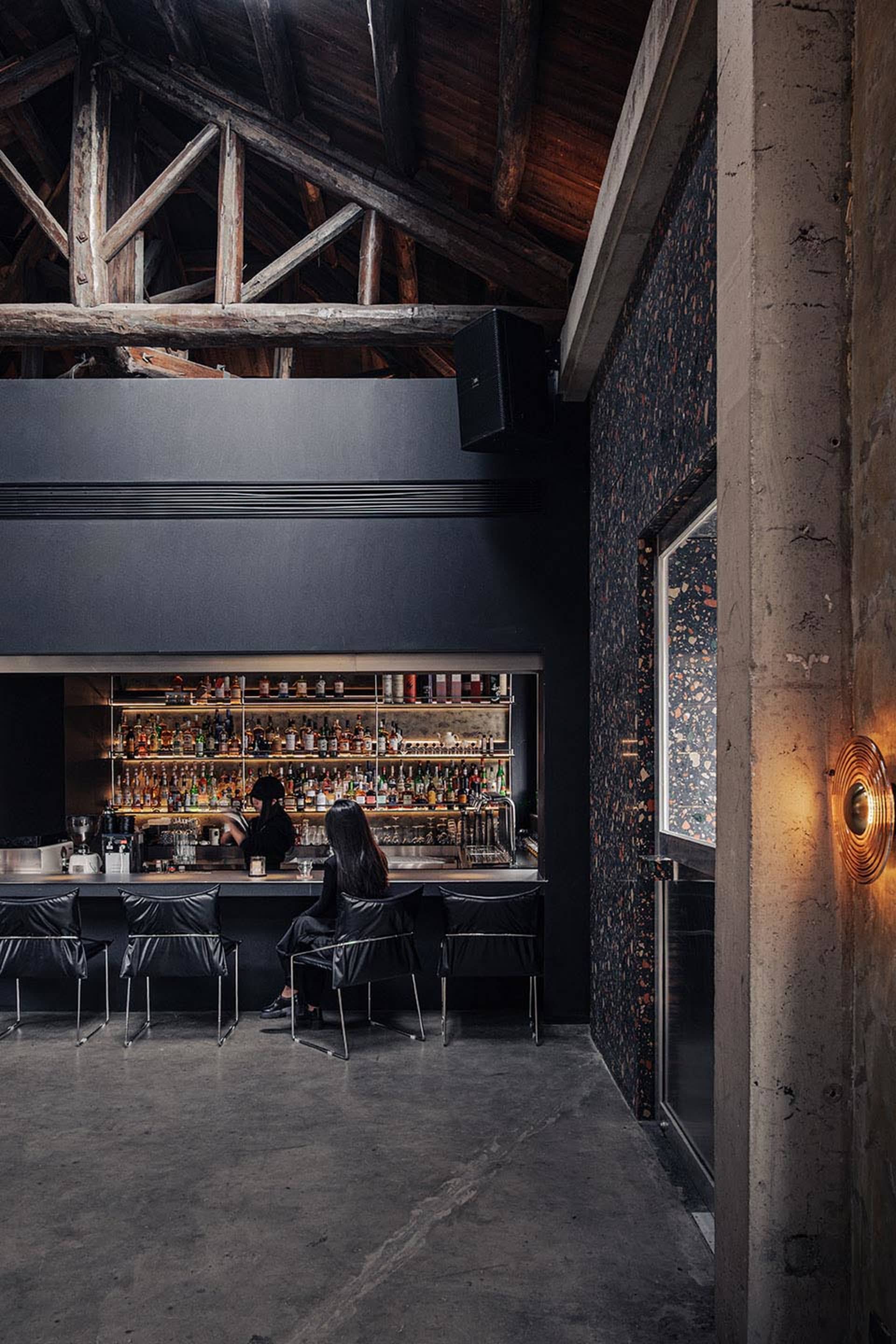 A casual bar scene with a black counter, bottles, and a woman sitting at the counter with chairs and a dining table in the background