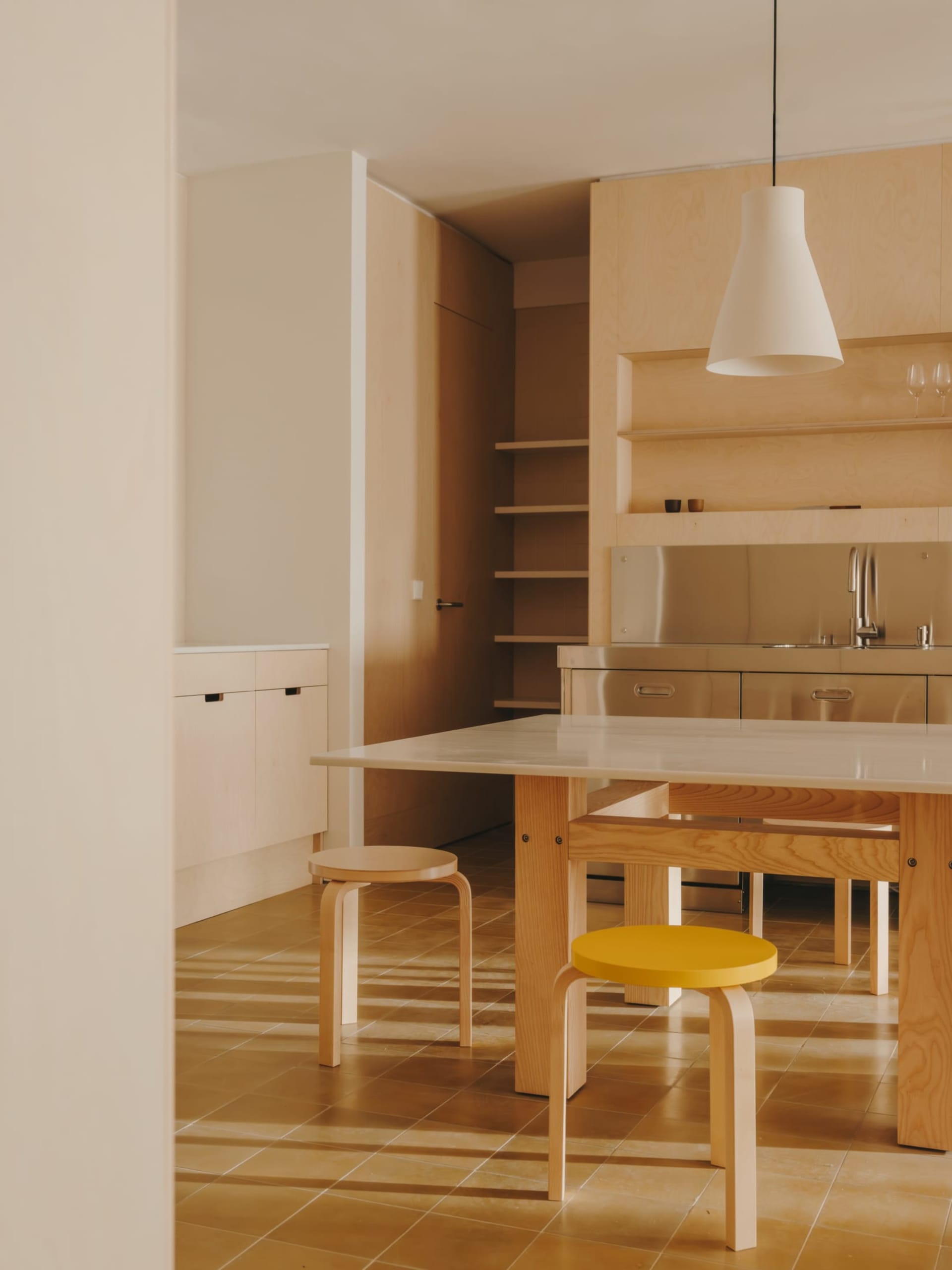 Modern kitchen with wooden floors and cabinets, featuring a sink, refrigerator, dining table, and chairs, with natural light and a warm atmosphere