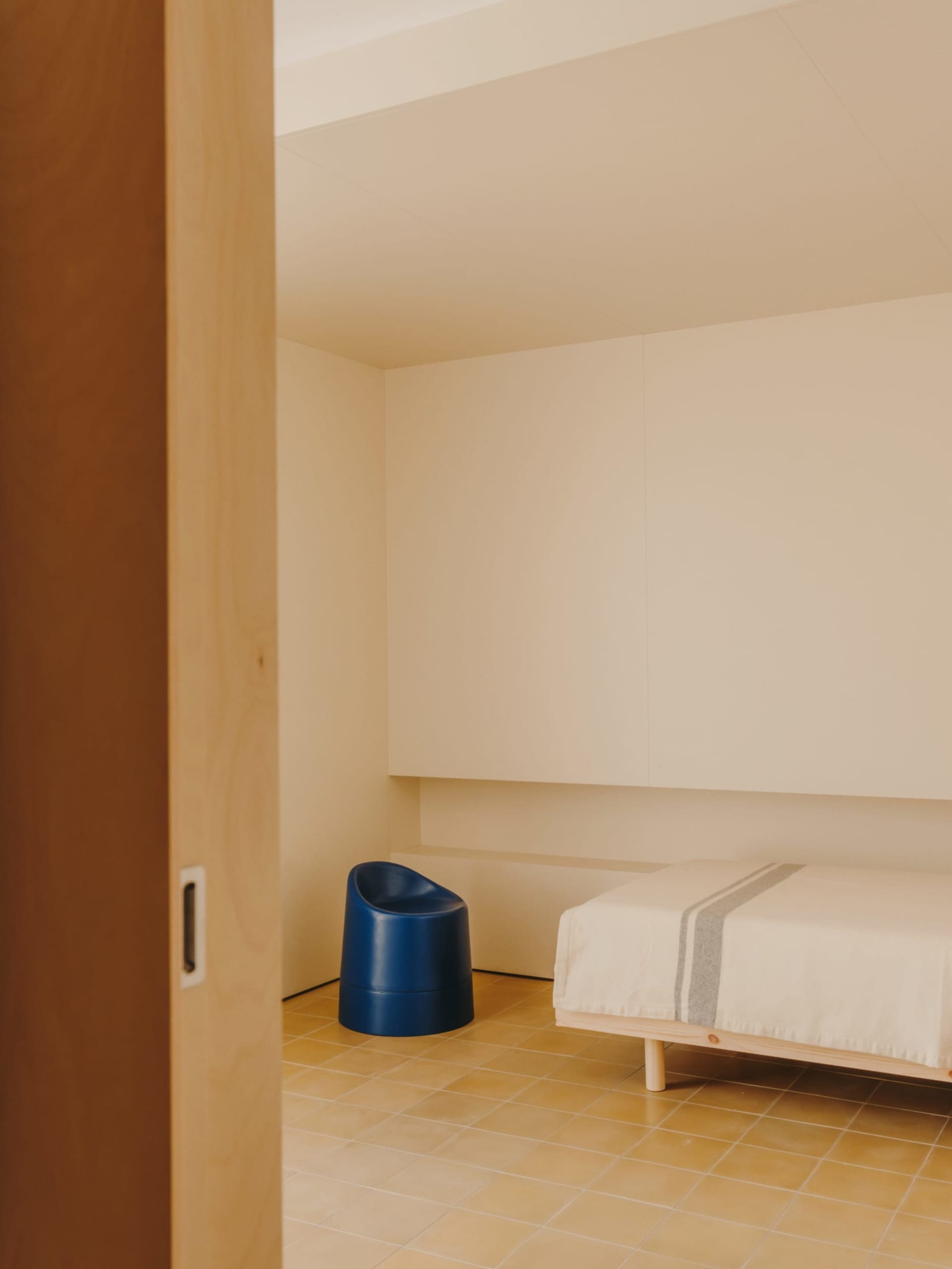 A small, simple bedroom with a bed against the wall, a chair nearby, and a blue trash can, featuring predominantly white colors and natural light from a window