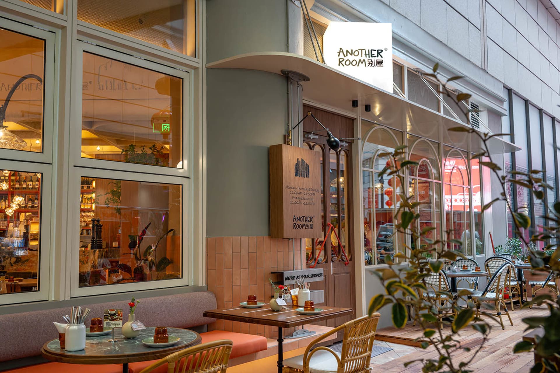 A dimly lit restaurant with a mix of wood and glass materials, featuring tables, chairs, and potted plants, creating a cozy and inviting atmosphere