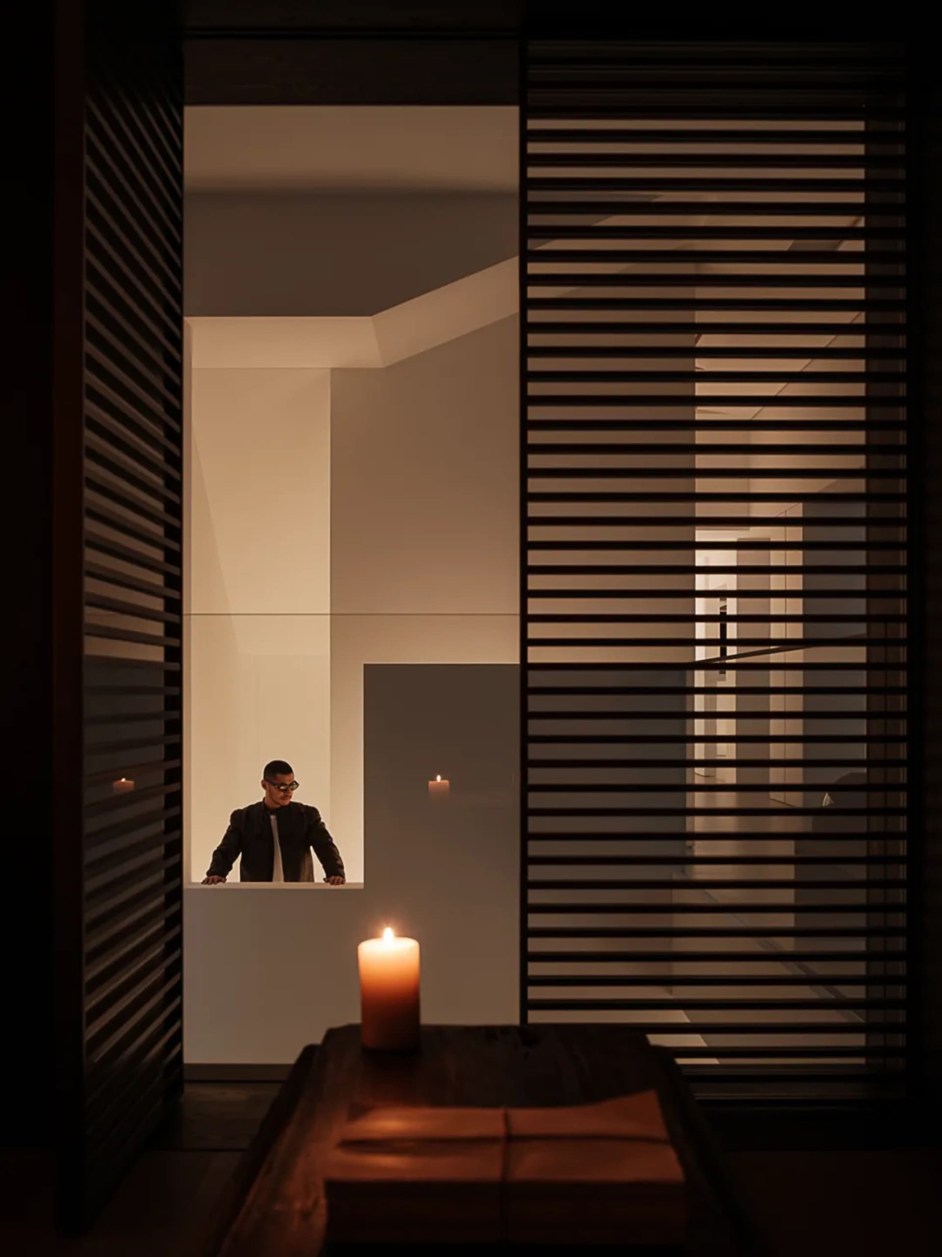 A man in a suit stands in a small room with a lit candle on a table, surrounded by a white wall and a doorway