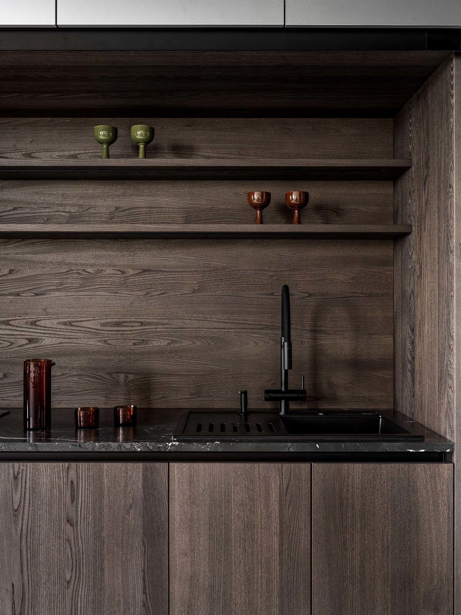 Modern kitchen with wooden cabinets, a black sink, and a wooden countertop, featuring a shelf with glasses and a dimly lit atmosphere