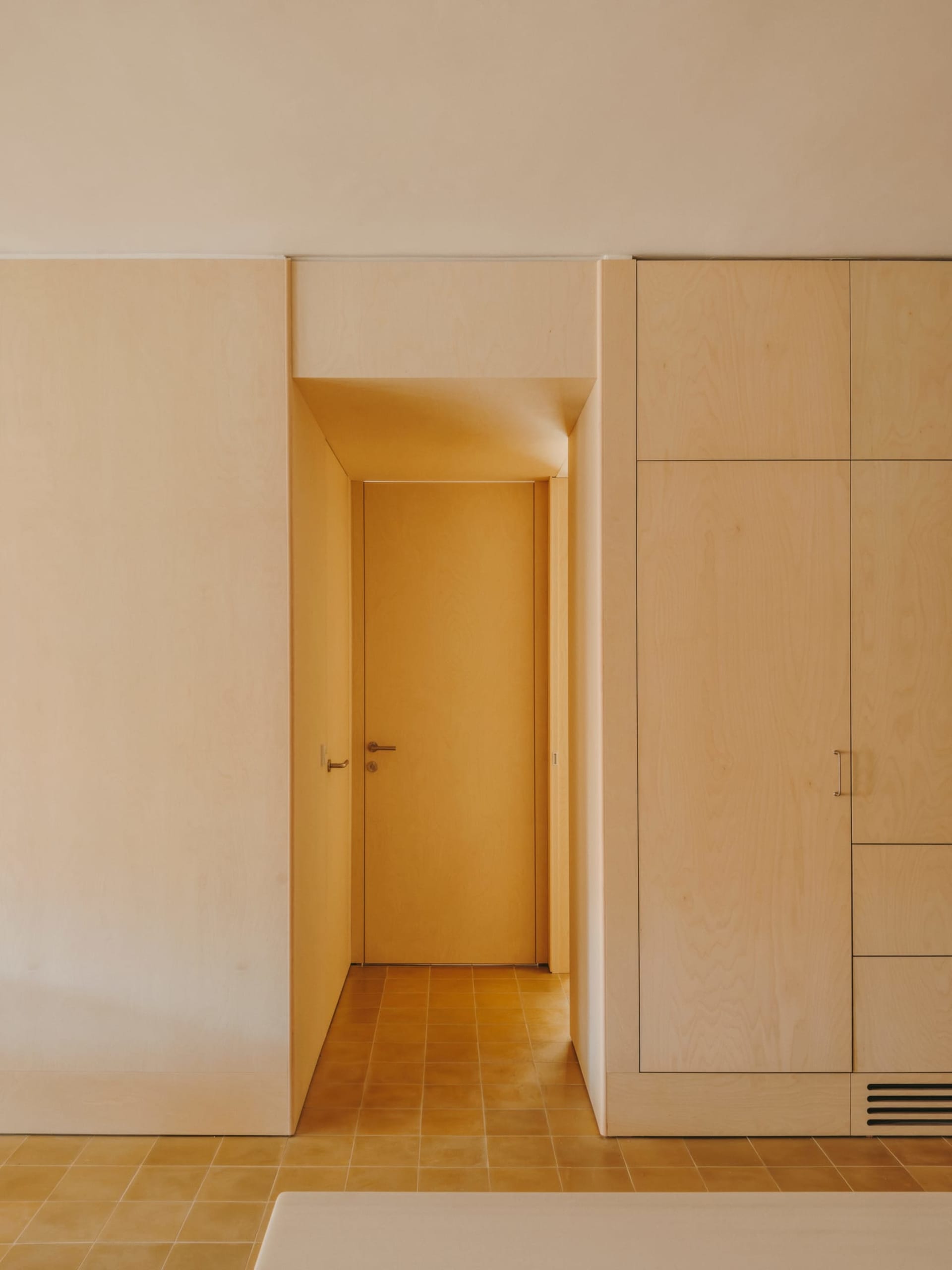 A calm and inviting bedroom with a white room, a brown door, and a tile floor, lit by natural light from the open doorway to the hallway