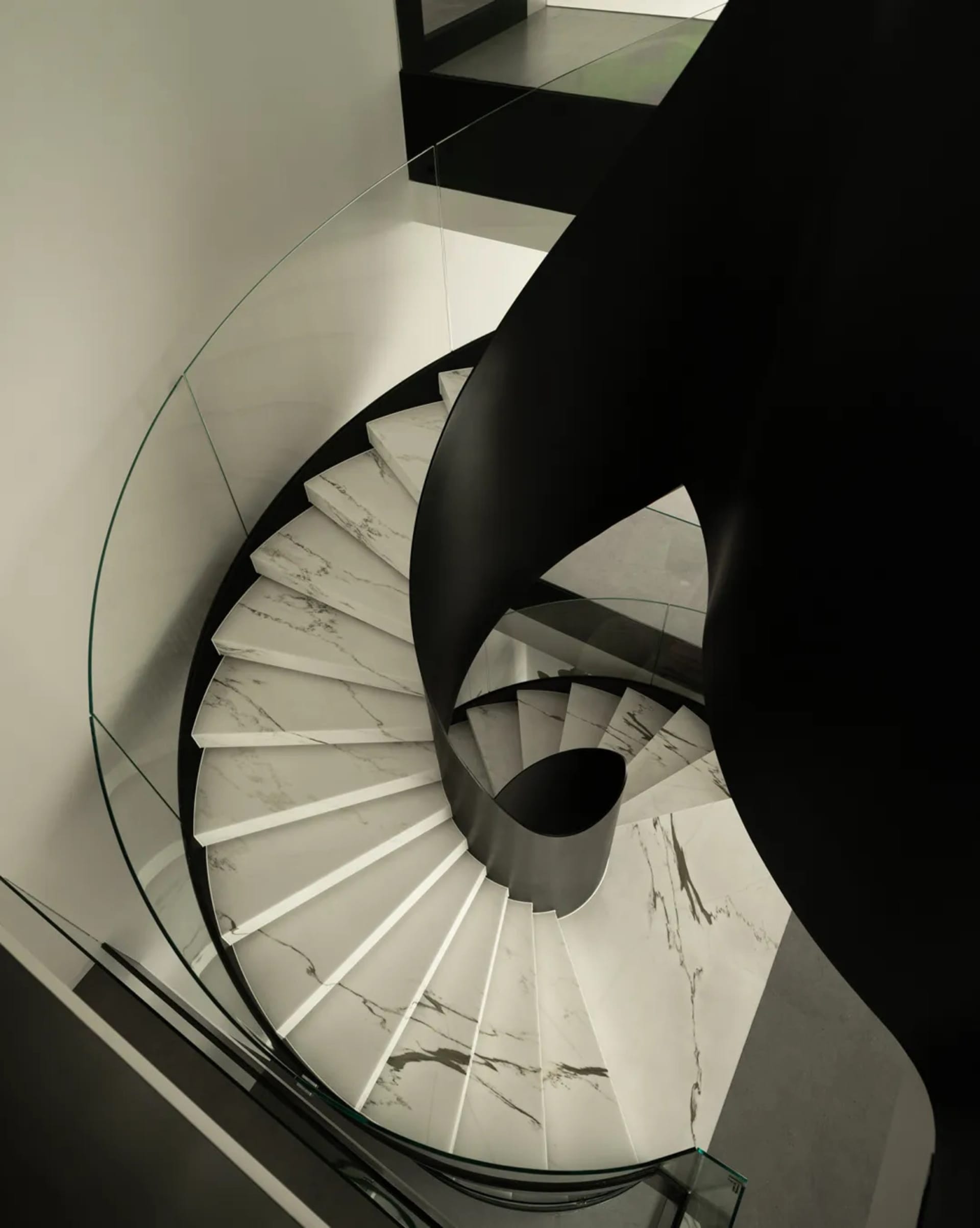 A black and white photograph of a spiral marble staircase in a room with a glass wall, surrounded by elegant decor and natural light