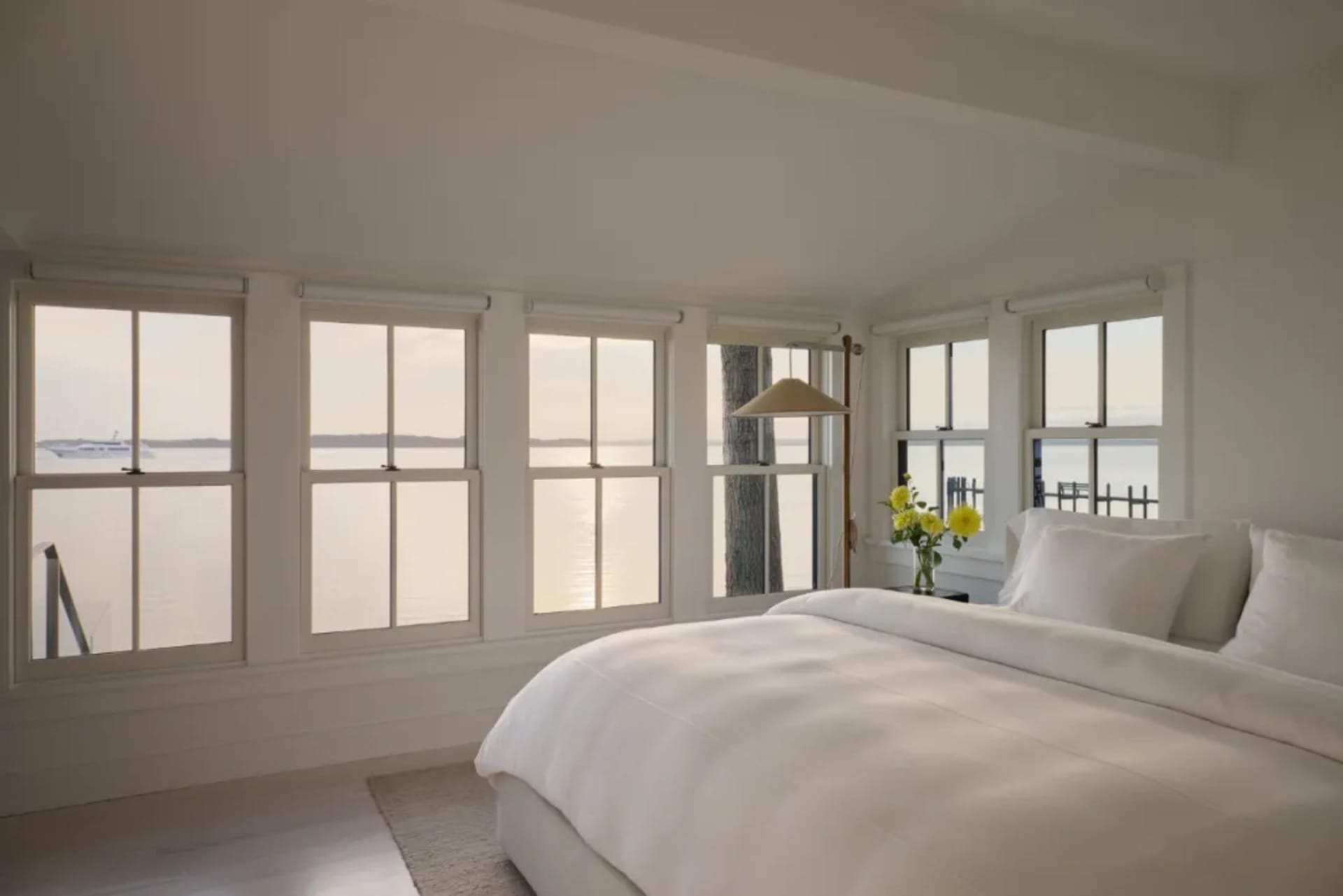A serene bedroom with a white comforter, yellow flowers, and a wooden floor, lit by natural sunlight through the window