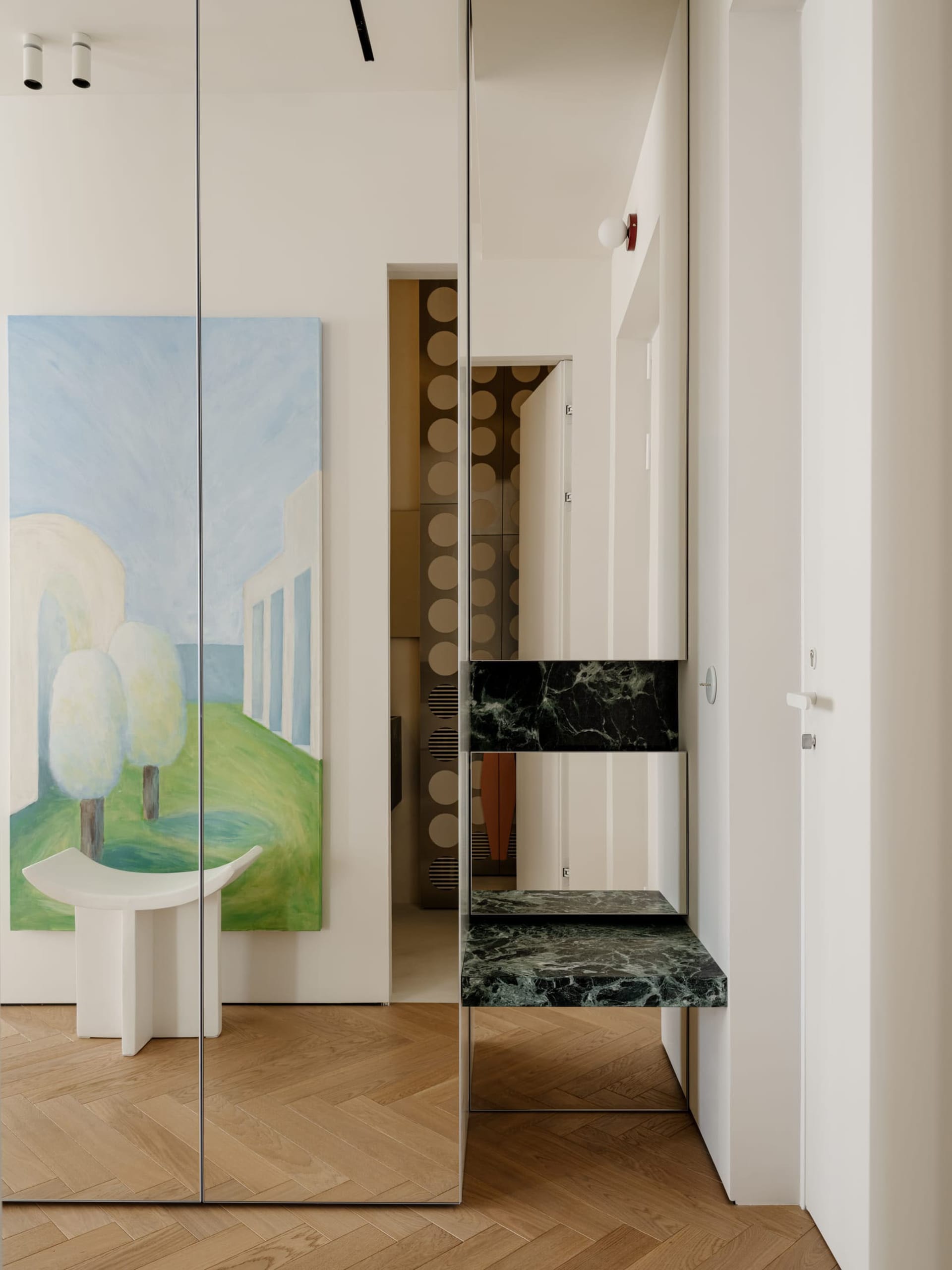 A modern living space with a white table, green painting, and mirrored wall, featuring a bench and chair in a clean and minimalistic design with natural light and white colors