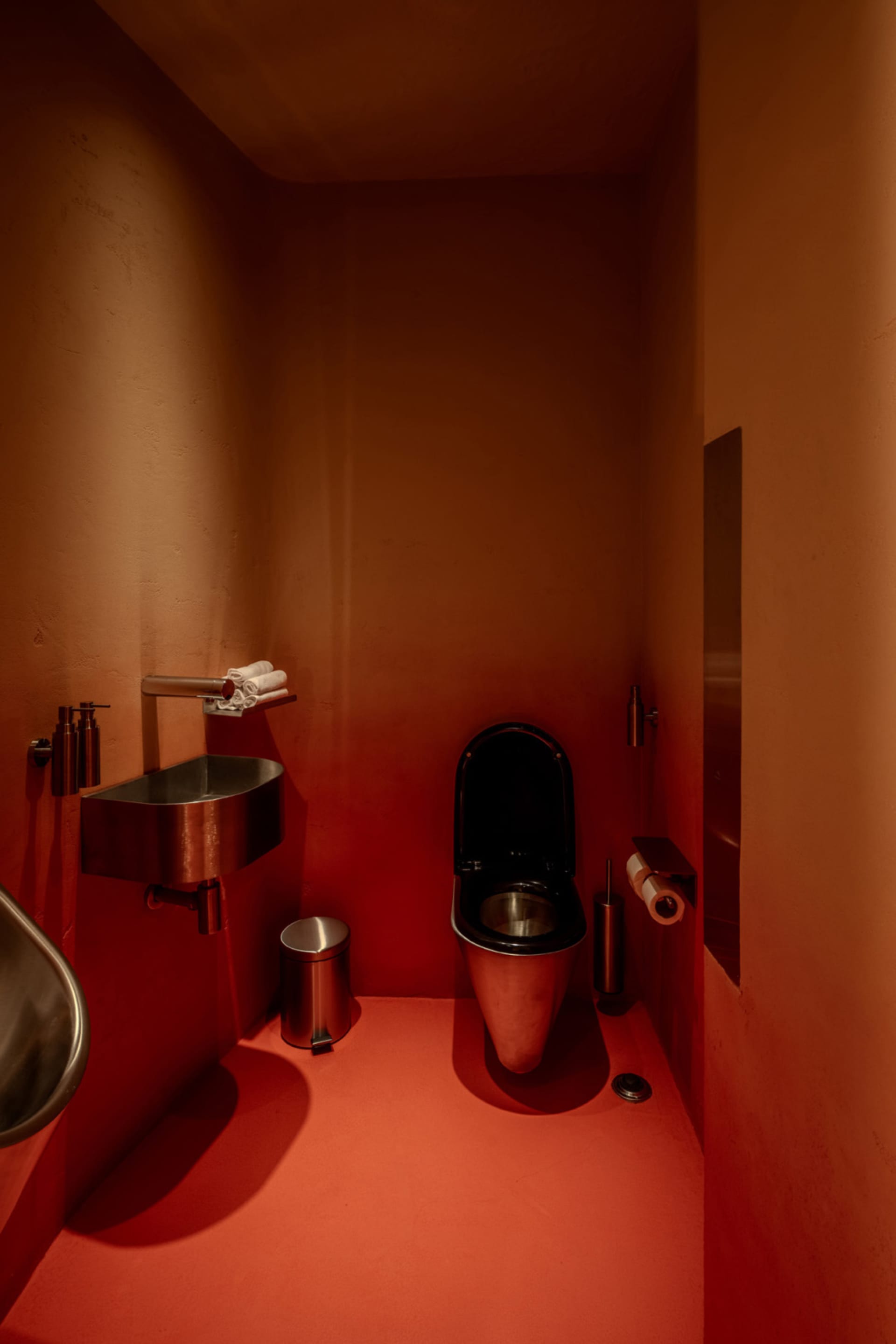 Bathroom with a black toilet, sink, and trash can, set against a bold red wall in a dimly lit space