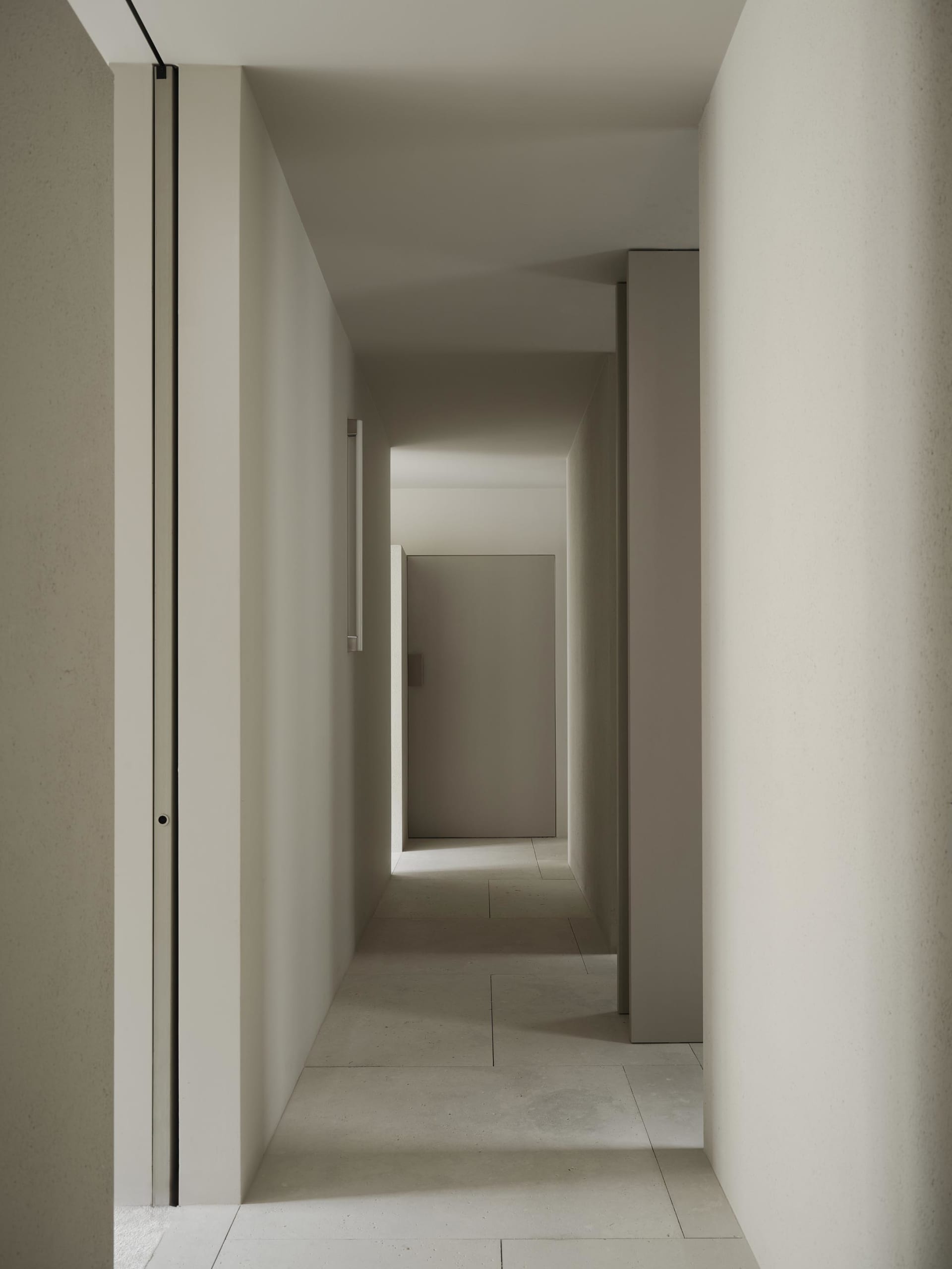 Narrow hallway with concrete walls, white color, and dim lighting, leading to an open doorway