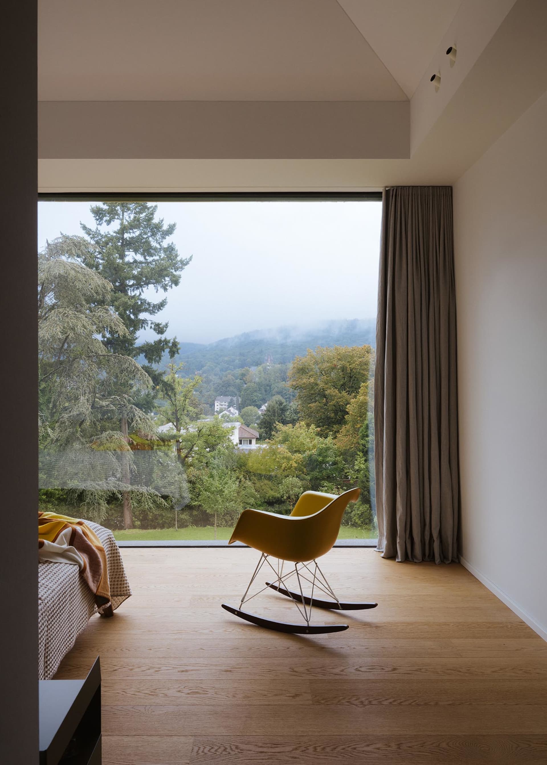 A modern living room with a bed, chair, and couch in front of a large window, surrounded by a warm and inviting atmosphere with natural light and wood materials