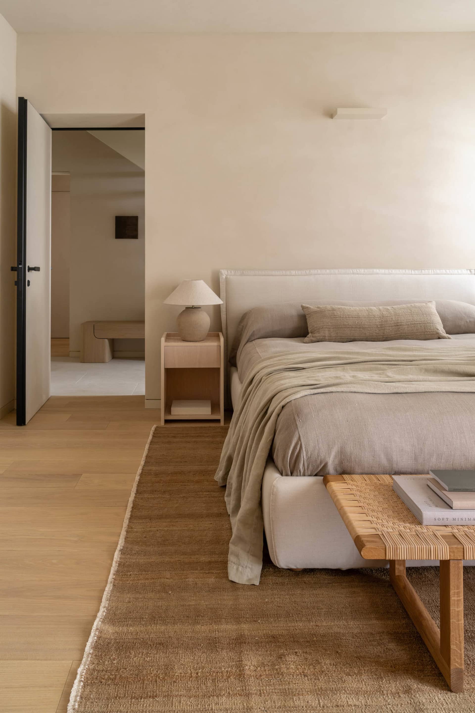 A modern bedroom with a wooden floor, a white wall, a bed with a white blanket and brown headboard, a table, and a chair, lit by natural sunlight