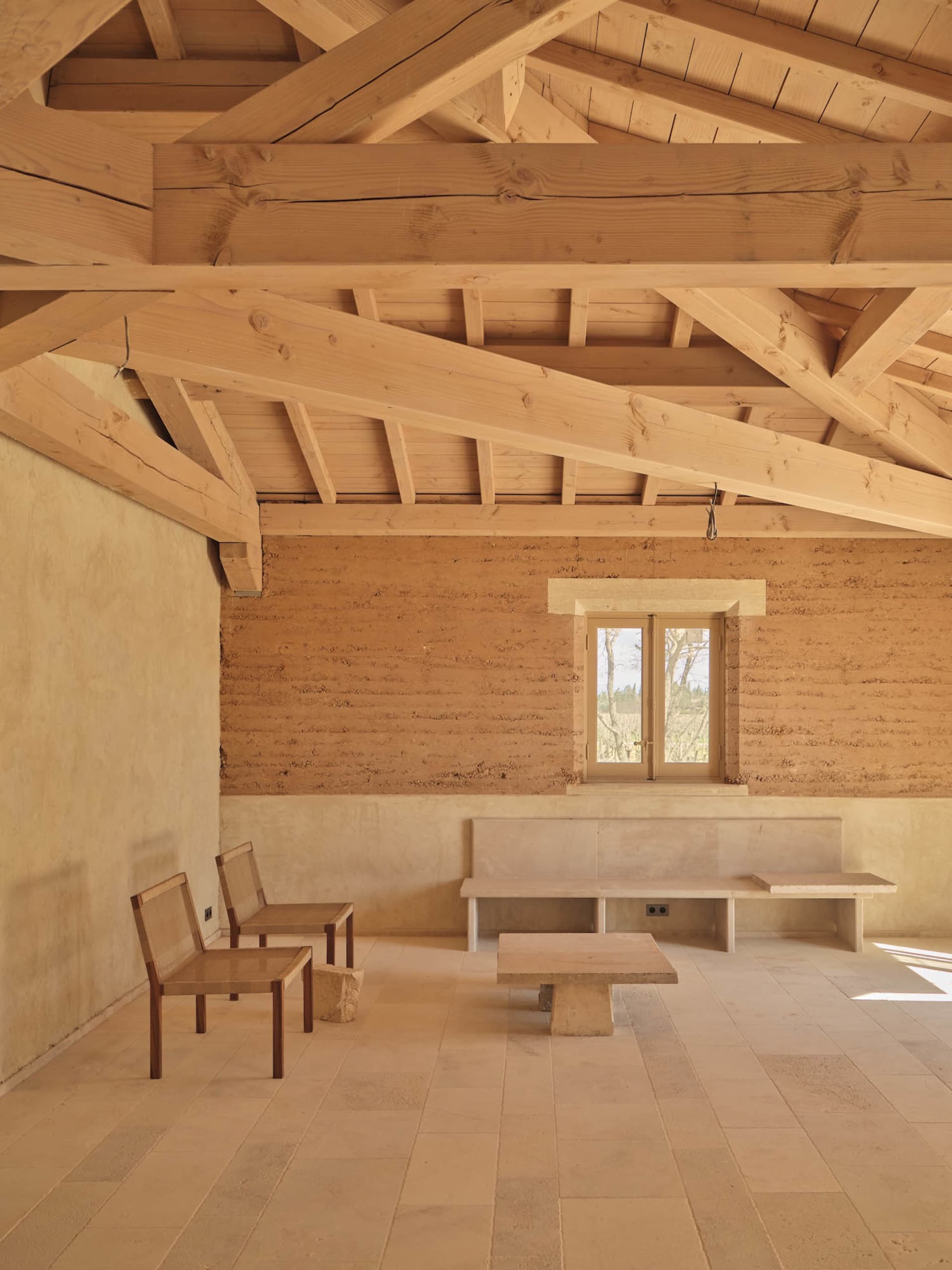 Dining area with a wooden floor, stone wall, dining table, chairs, and bench, lit by natural sunlight through a window