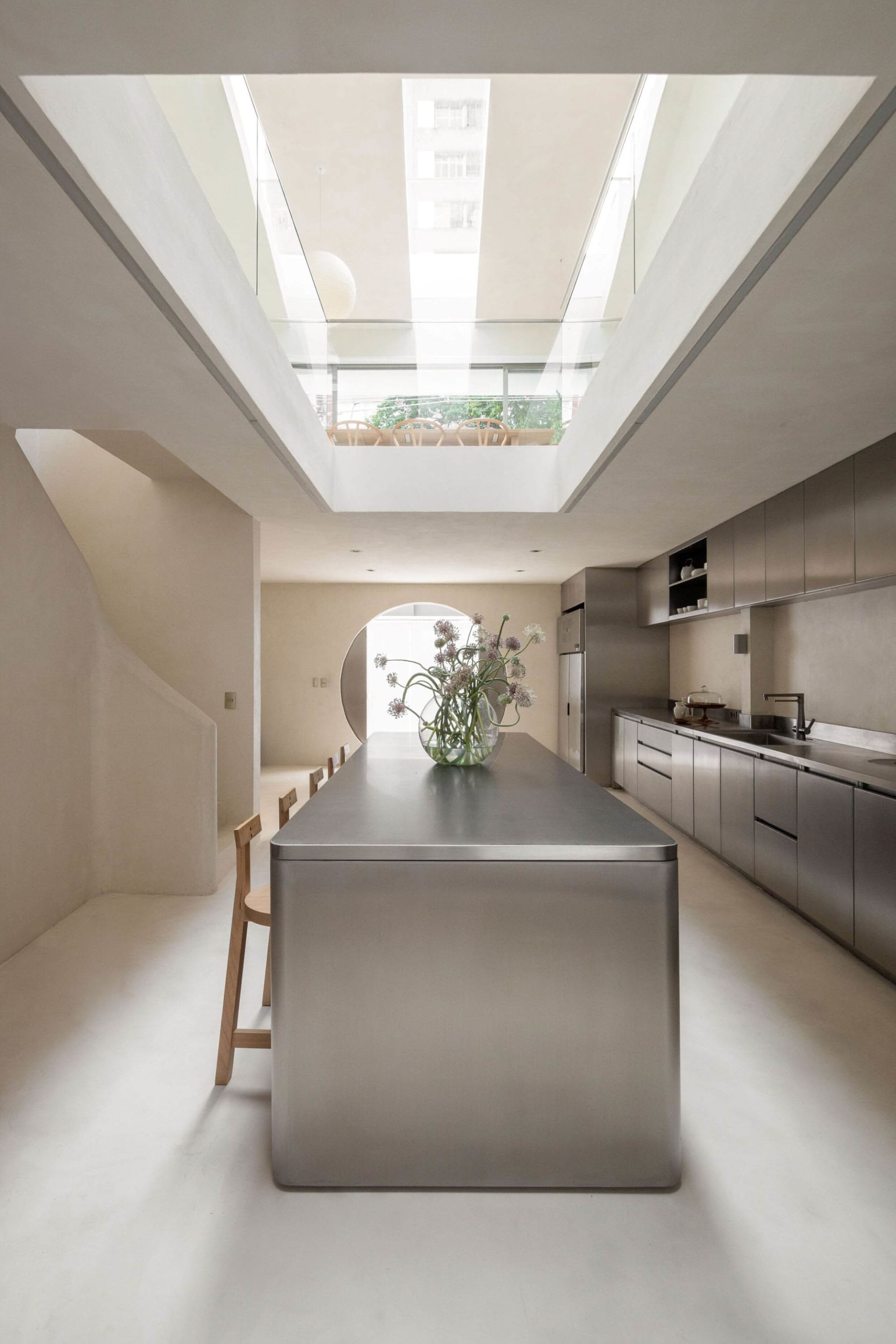 Modern kitchen with a stainless steel island, a dining table, and a refrigerator, featuring a minimalist and well-lit space with a vase with flowers on top of the island