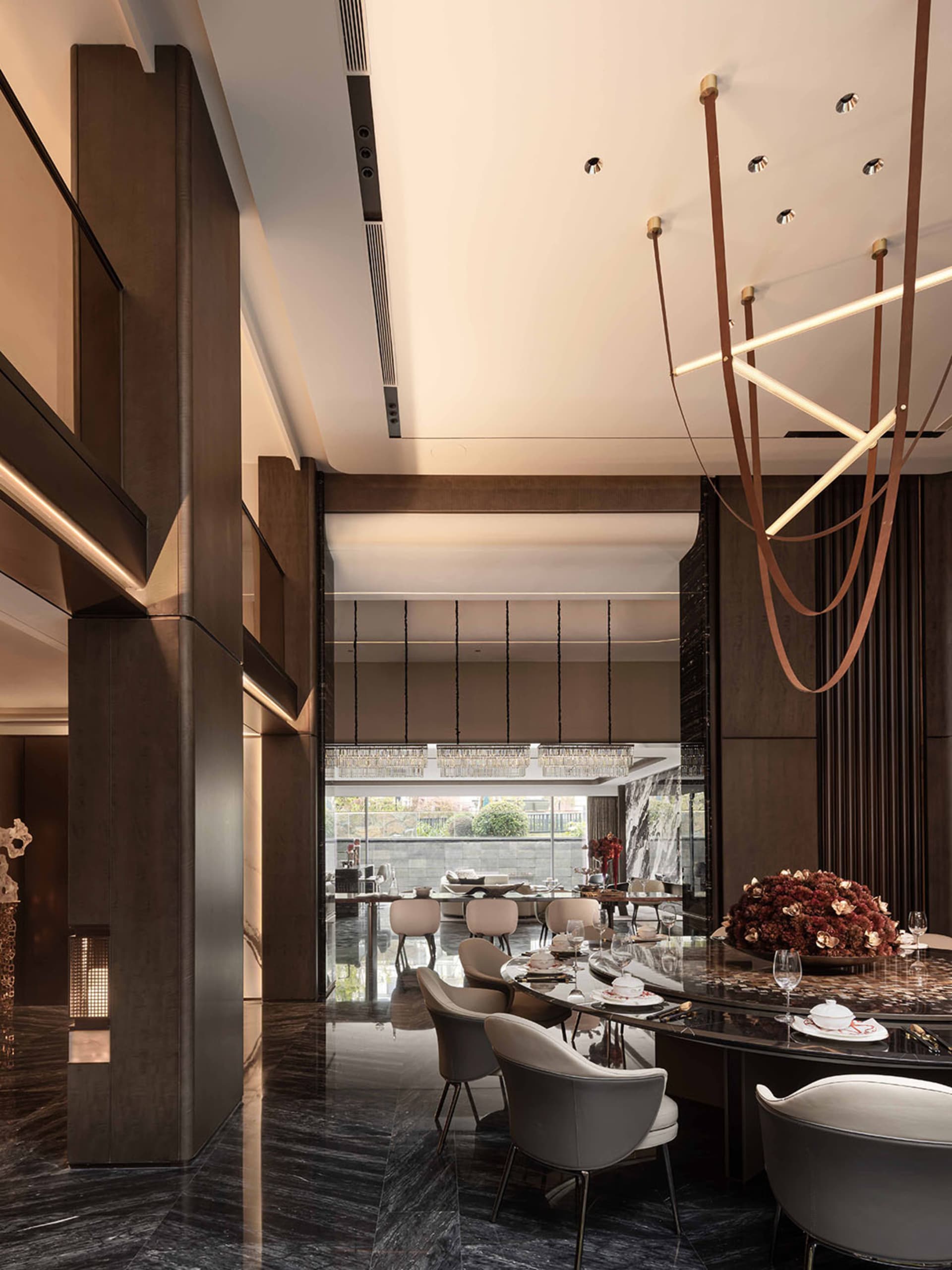 Spacious modern restaurant dining area with a large wooden table, chairs, and dim lighting, set for a meal with wine glasses, cups, and bowls